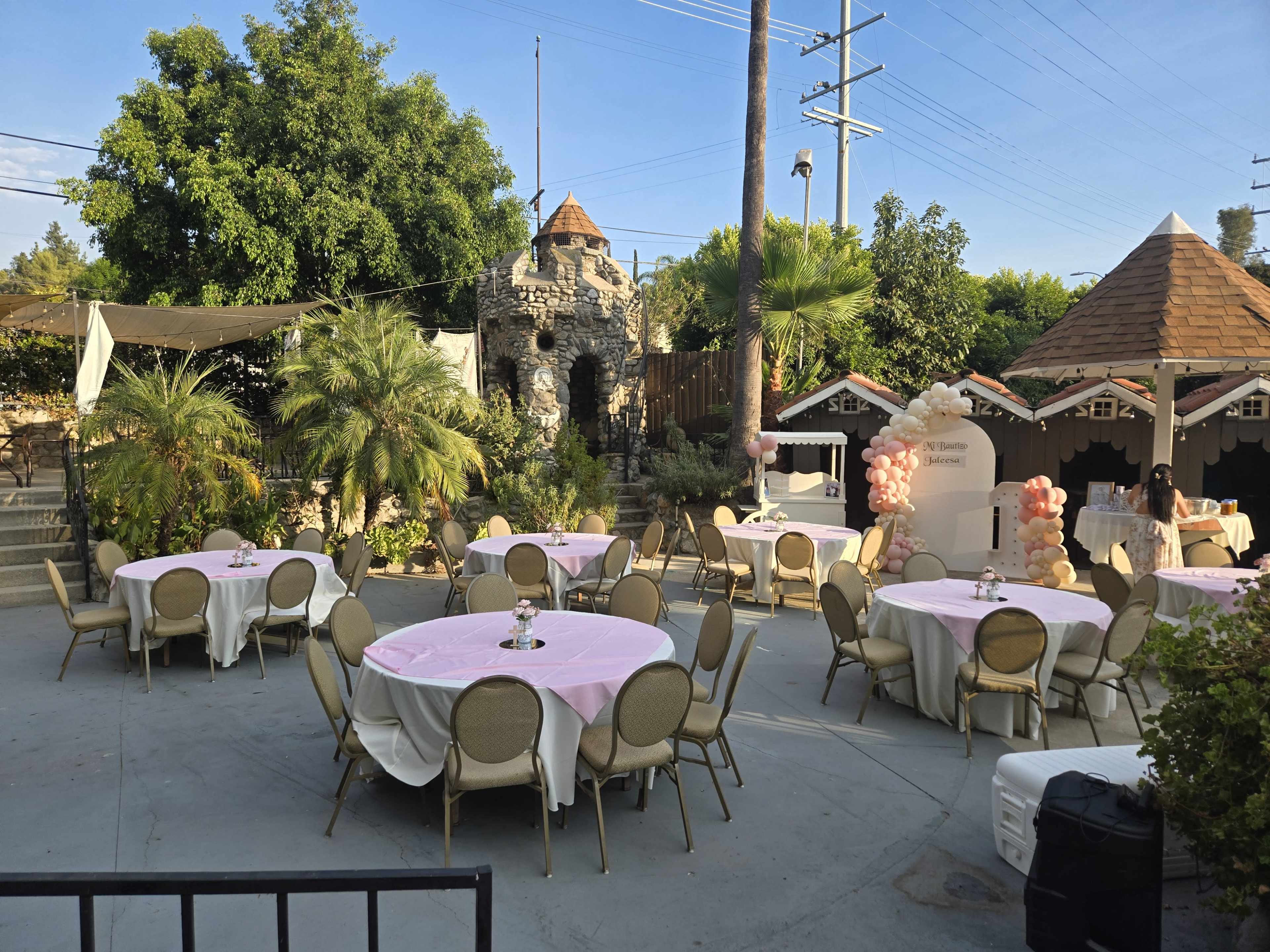 The image shows an outdoor event space with round tables covered in pink tablecloths, surrounded by greenery and a stone structure in the background.