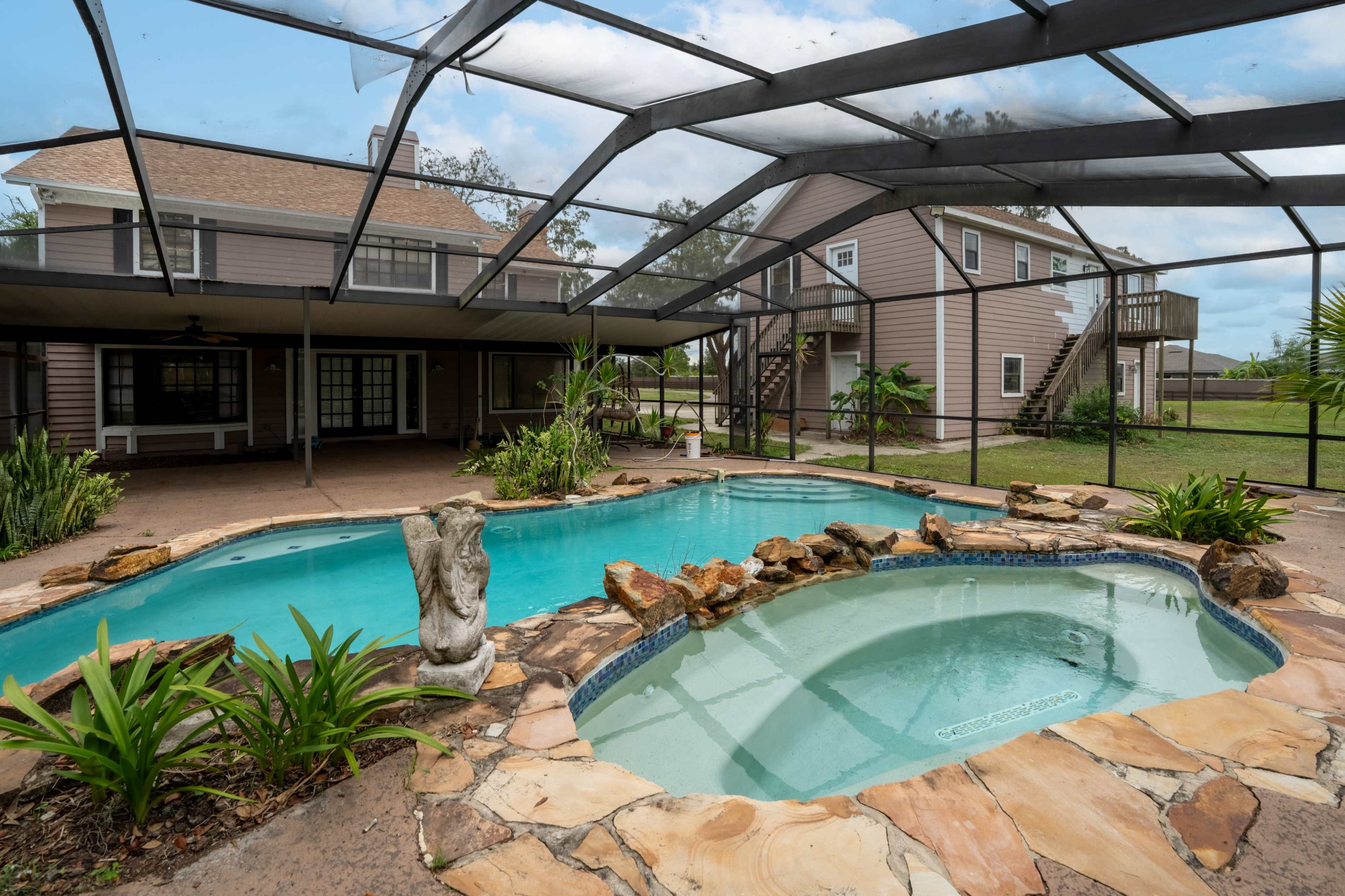 The image shows a spacious outdoor area with a swimming pool and a hot tub, surrounded by a screened enclosure and a two-story house in the background.
