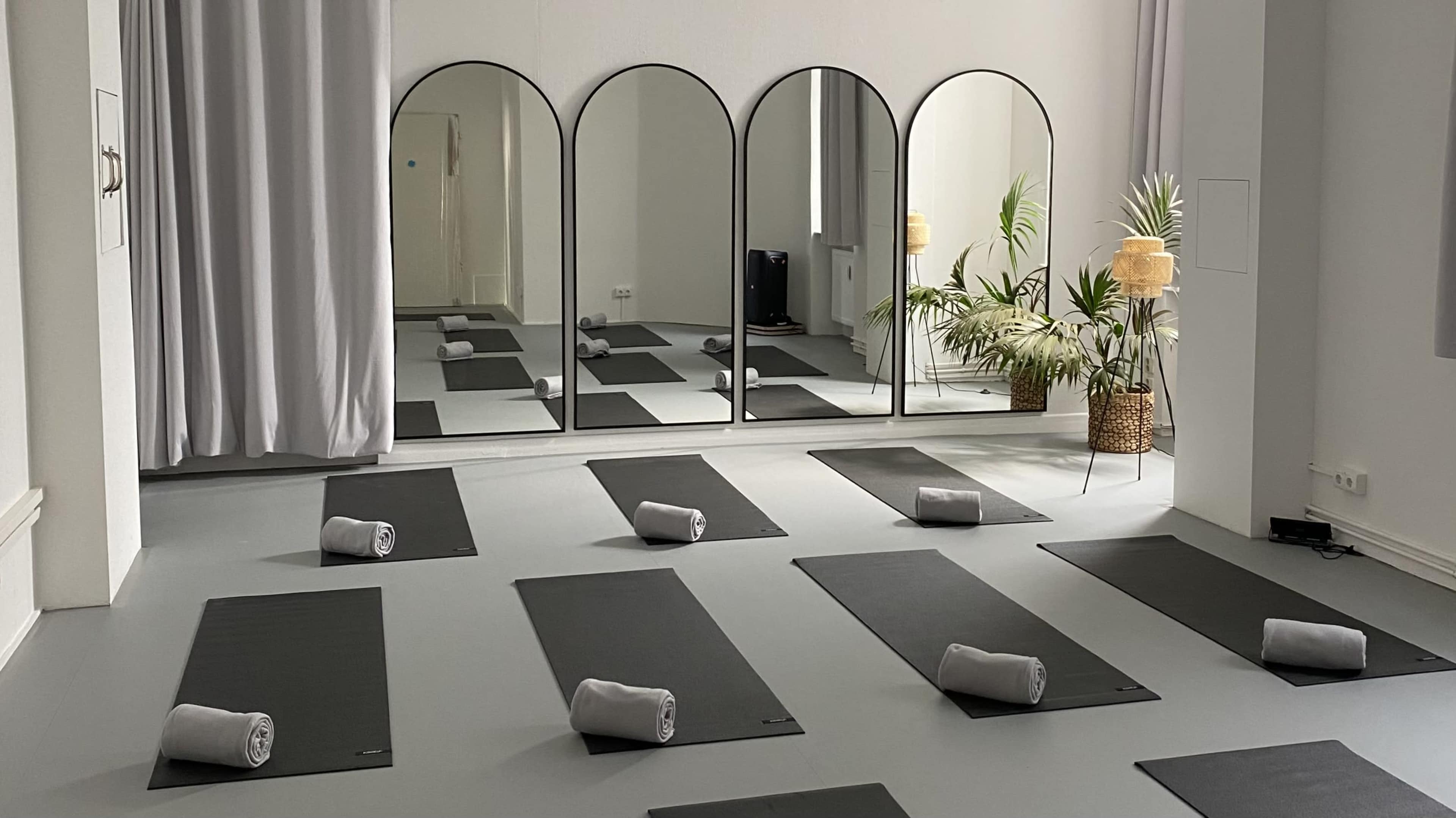 A yoga studio features multiple black mats arranged on the floor, each with a rolled towel, while large mirrors reflect the space and decorative plants are positioned in the corners.