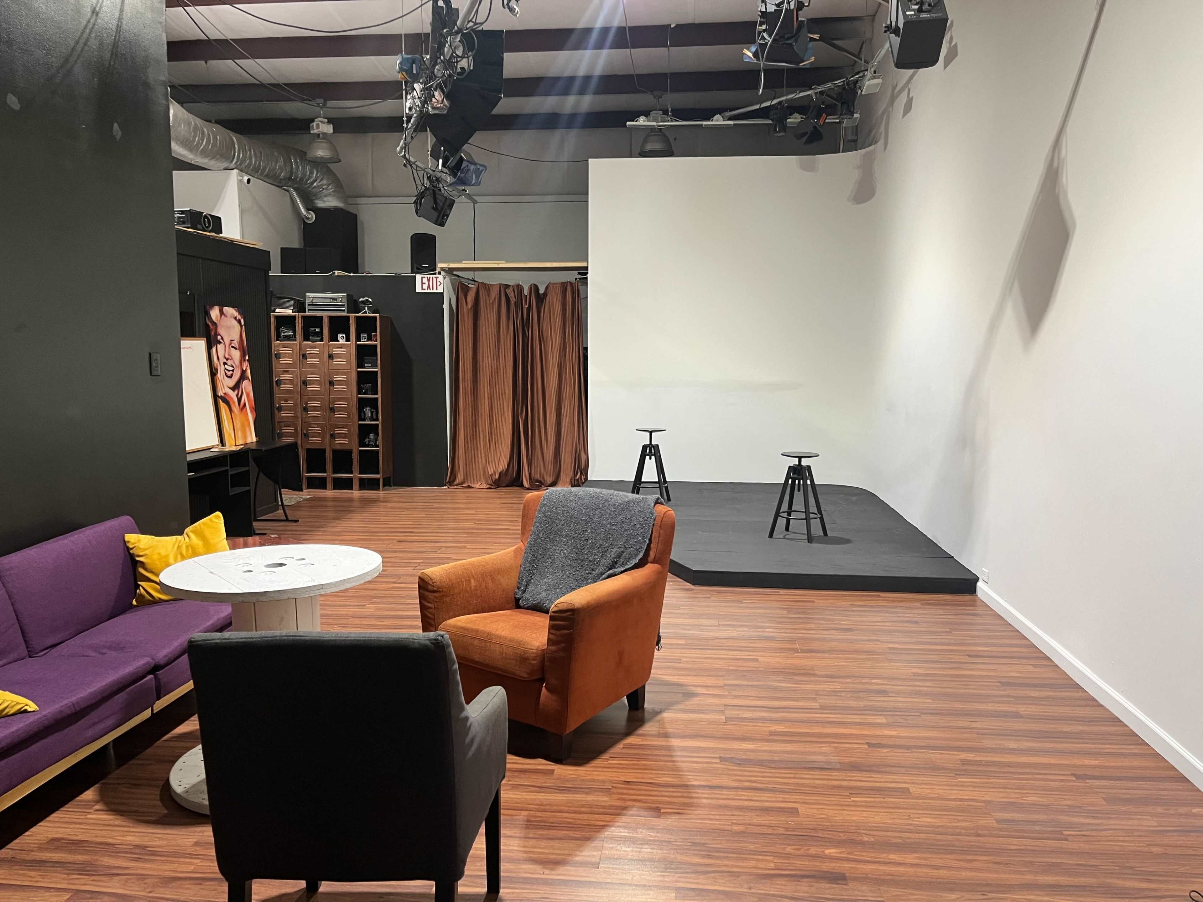 The image shows a modern studio space with a seating area featuring a purple couch, an orange armchair, and a round table, alongside a performance stage with two stools and a backdrop.