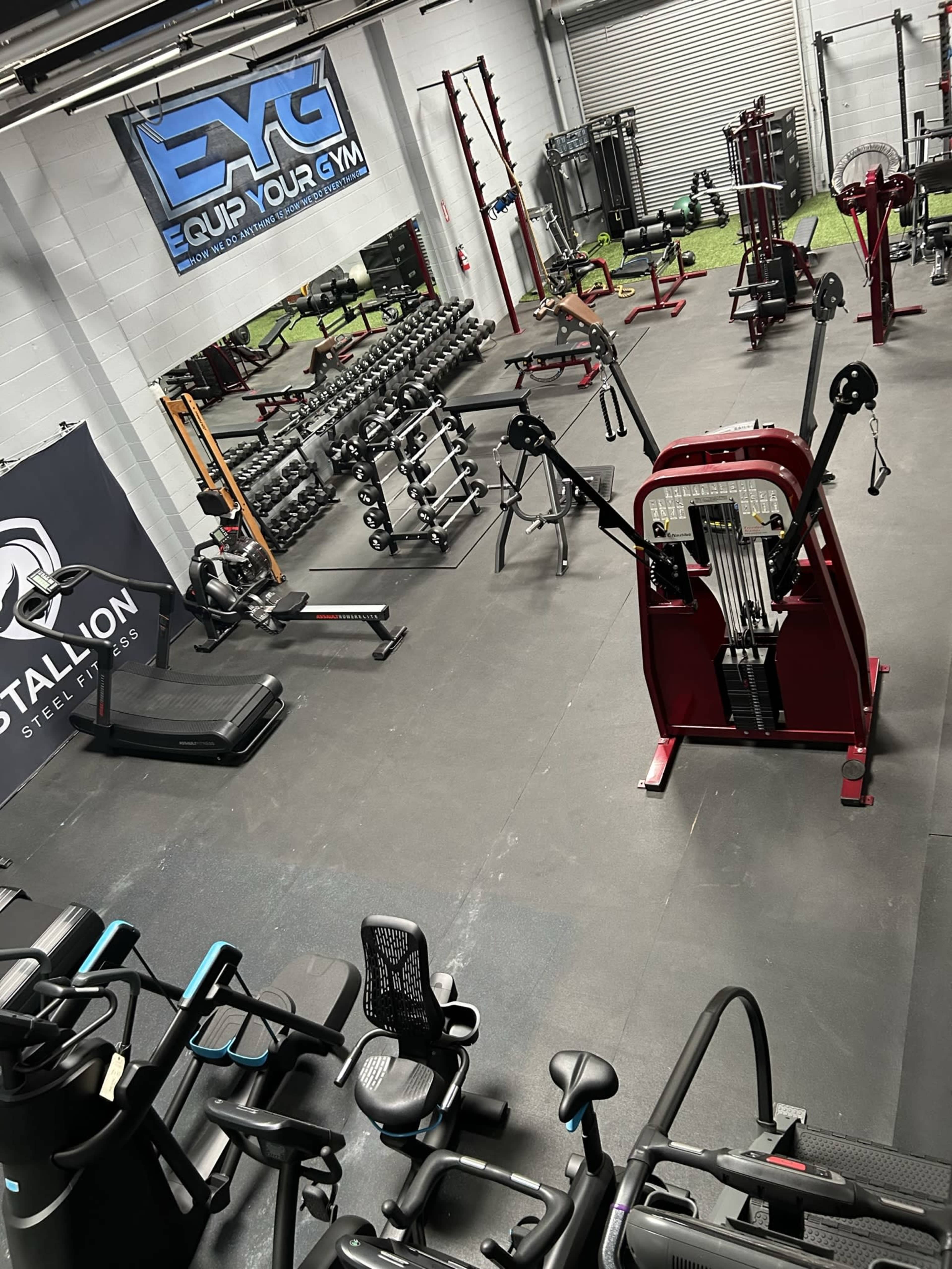 A workout facility equipped with various exercise machines, free weights, and gym equipment arranged on a rubberized floor.