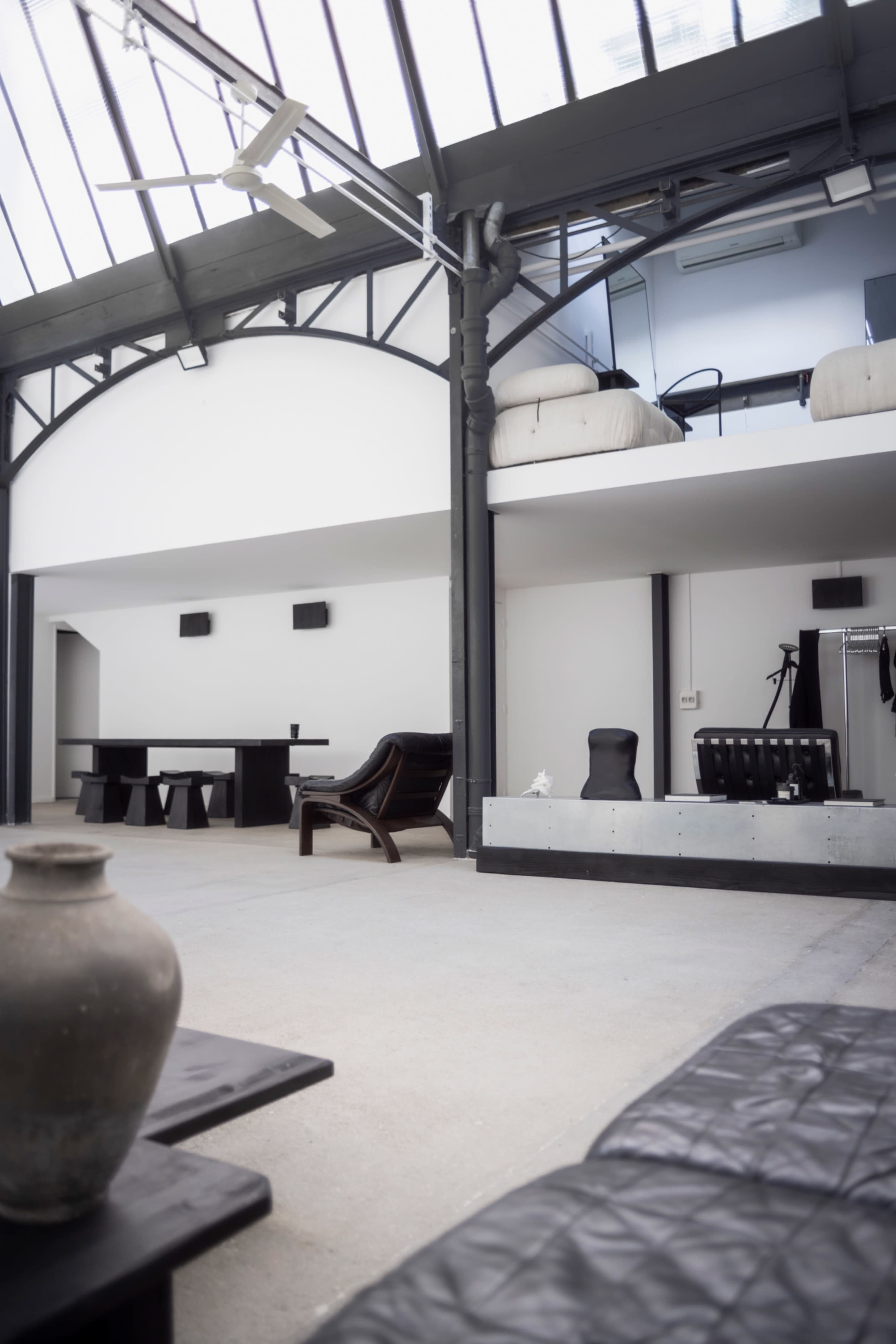 The image shows a spacious, industrial-style interior with high ceilings, featuring a mix of furniture including a wooden table, a lounge chair, and a minimalist bed area on a loft.