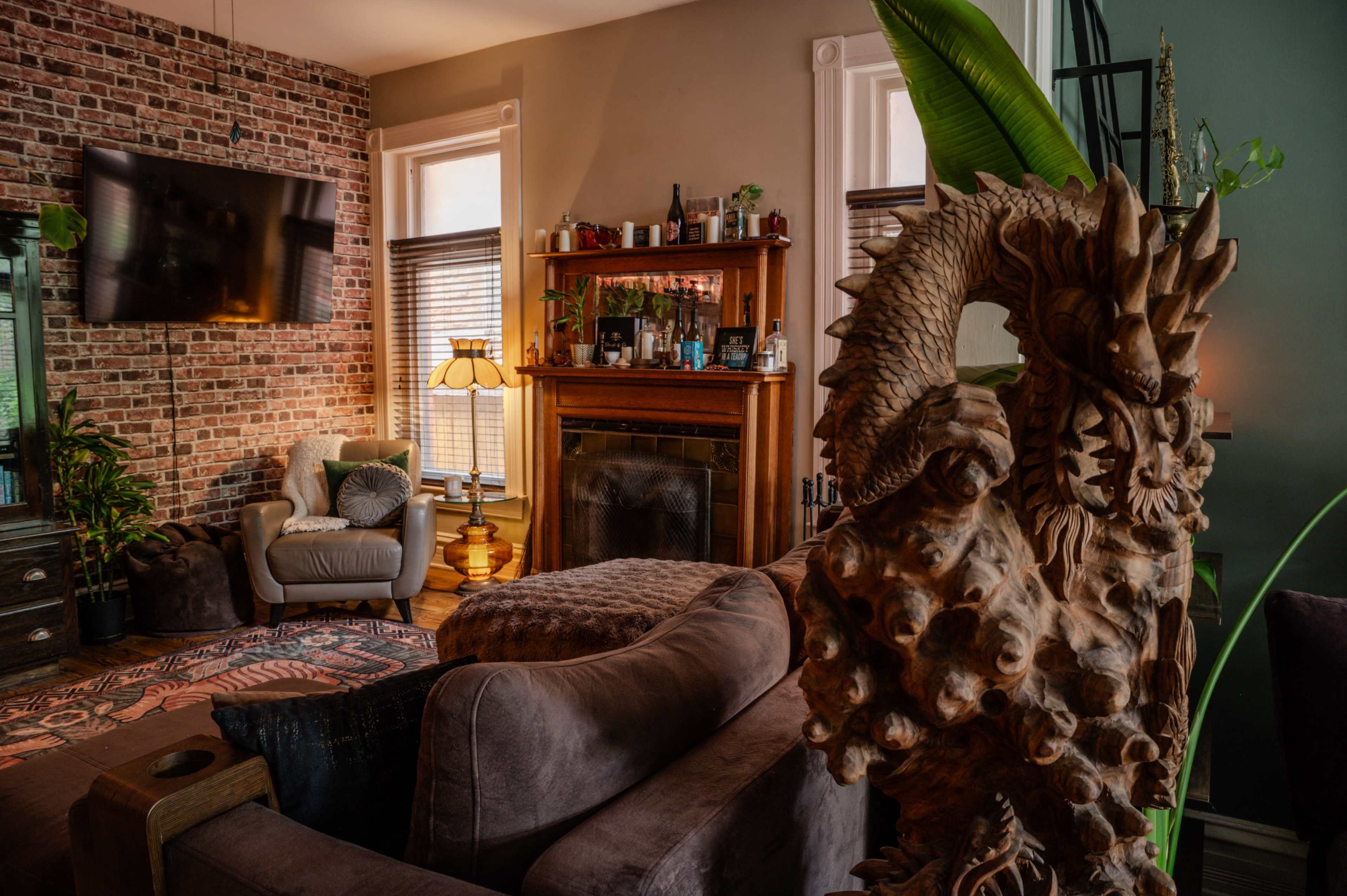 The image shows a cozy living room with a large TV mounted on a brick wall, a wooden shelf filled with bottles, and a carved wooden sculpture prominently displayed in the foreground.