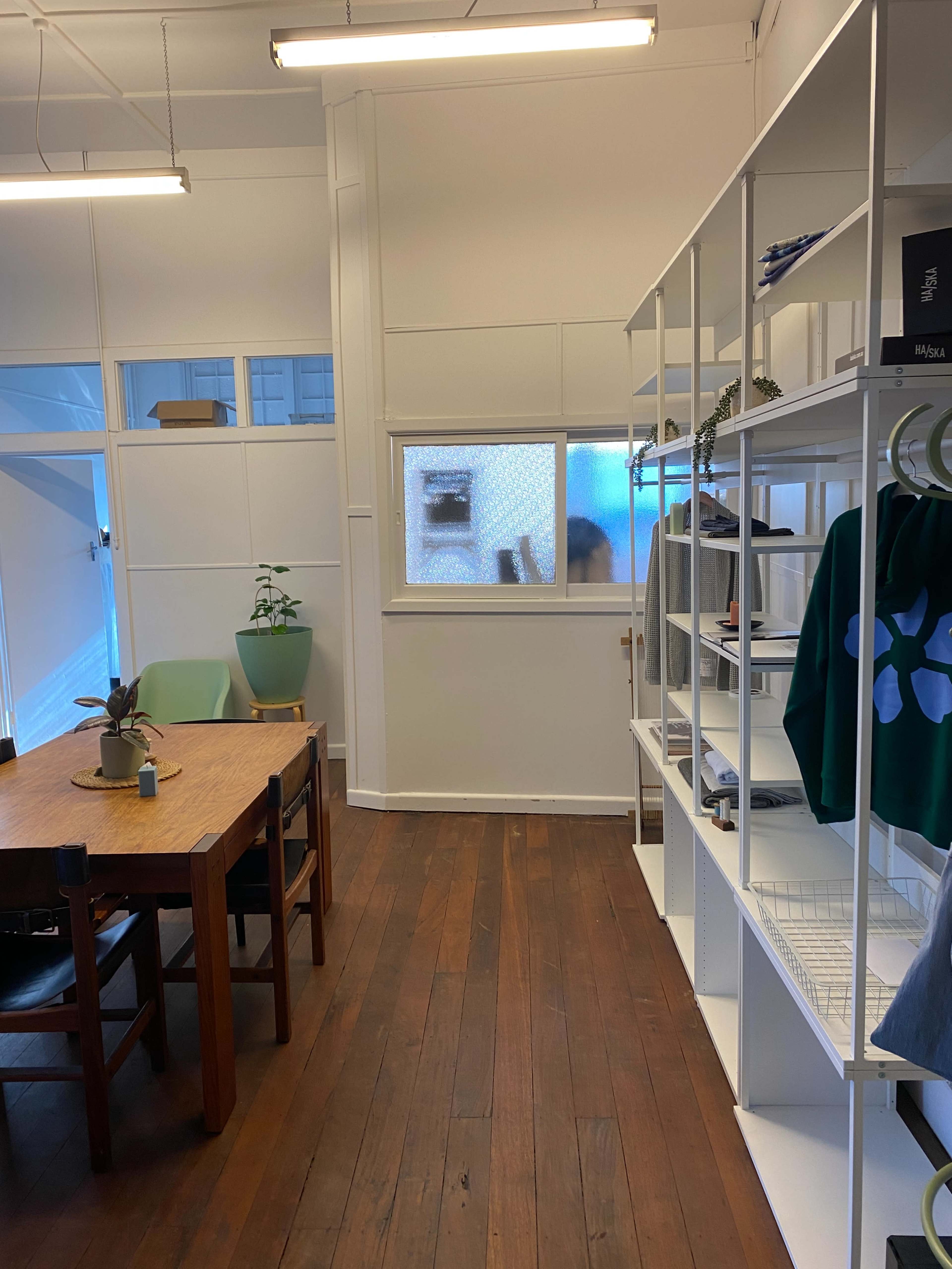 The image shows a well-lit room with wooden flooring, featuring a table with chairs, a plant, and a shelving unit displaying various items.