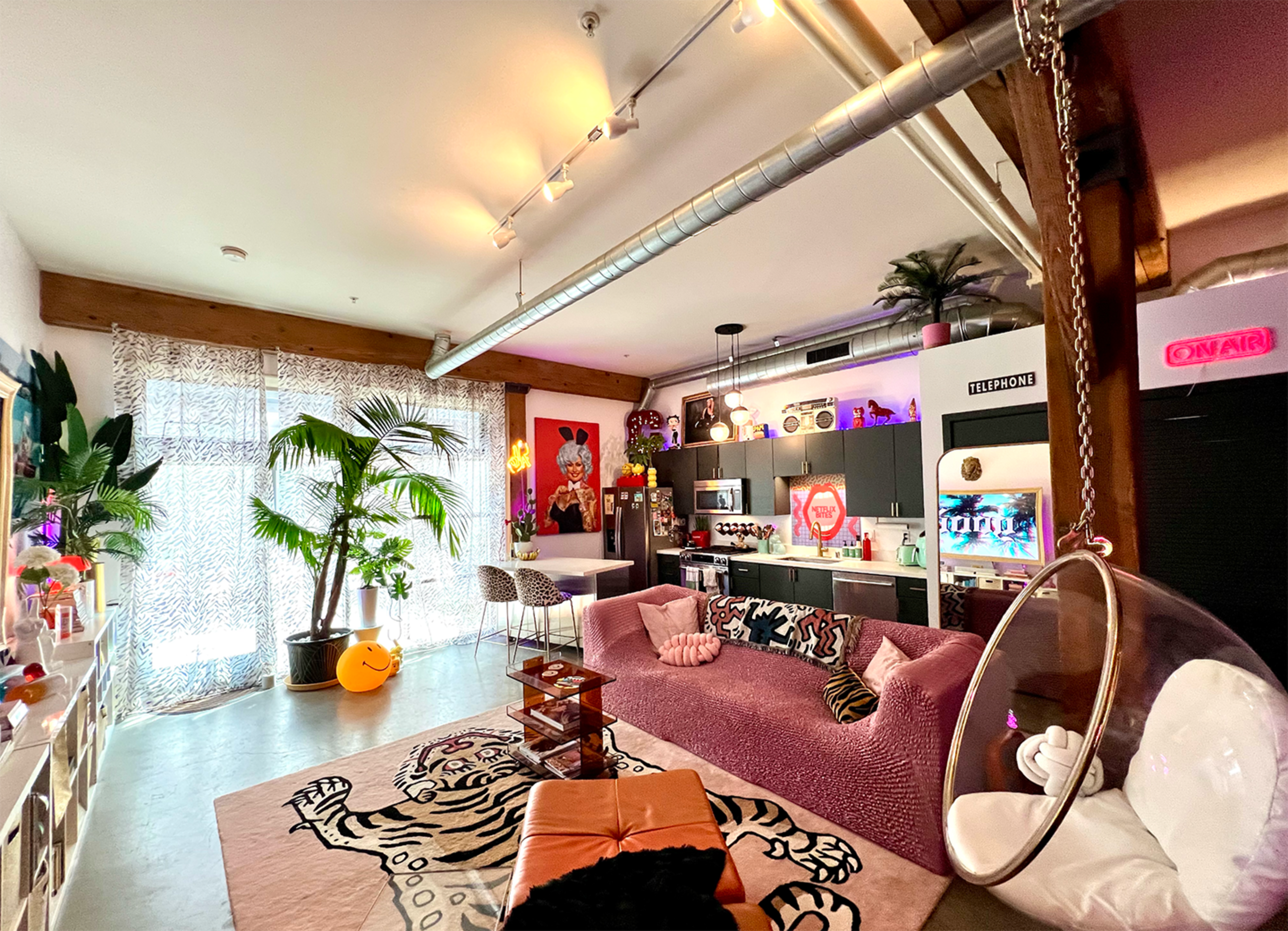A brightly lit, eclectic living room features a pink sofa, a large patterned rug, various decorative plants, and colorful art pieces on the walls, alongside a modern kitchen area.