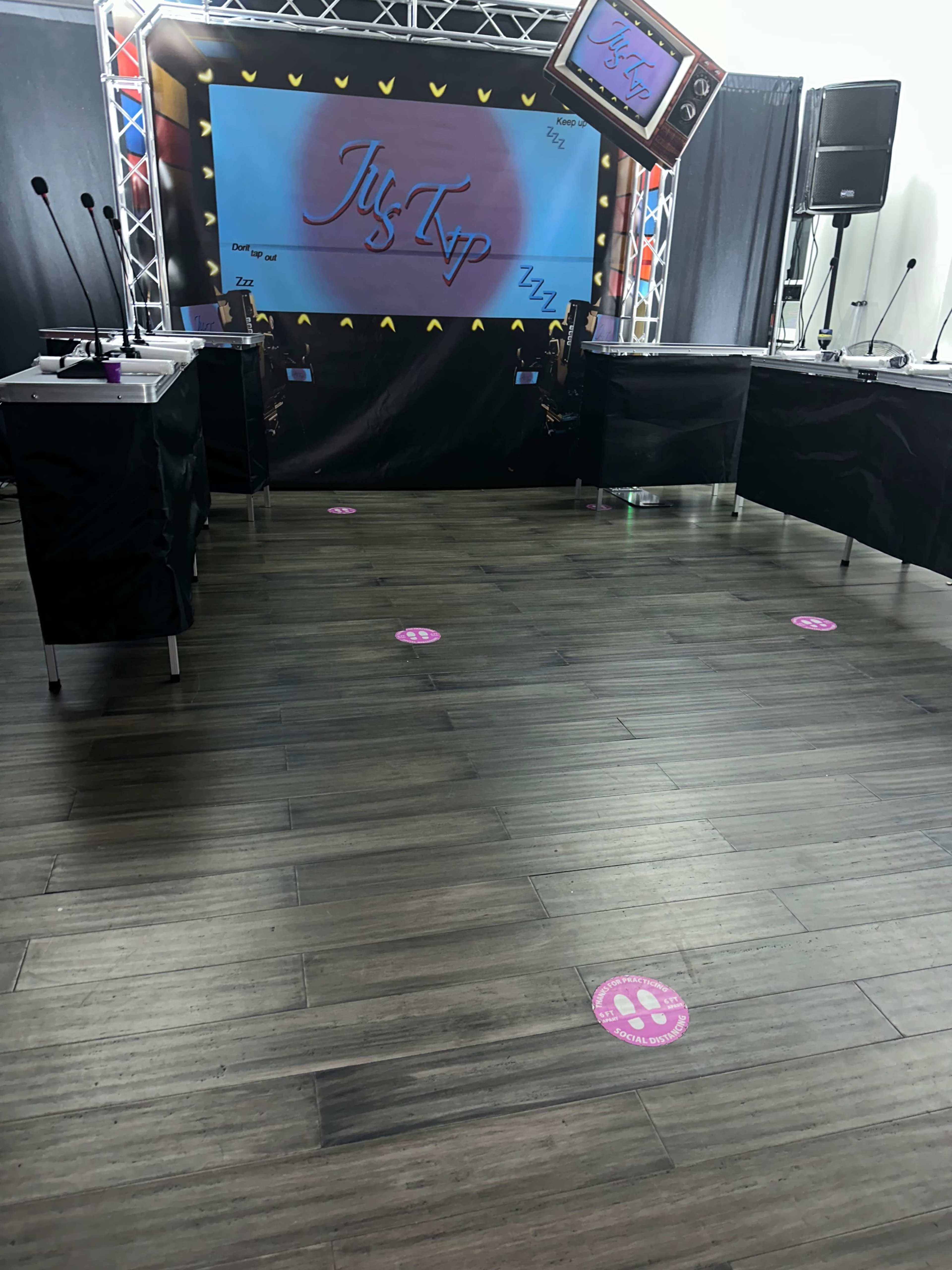 The image shows a rectangular room set up for an event, featuring a stage with a large screen and several tables with microphones, along with pink paw print markers on the floor.