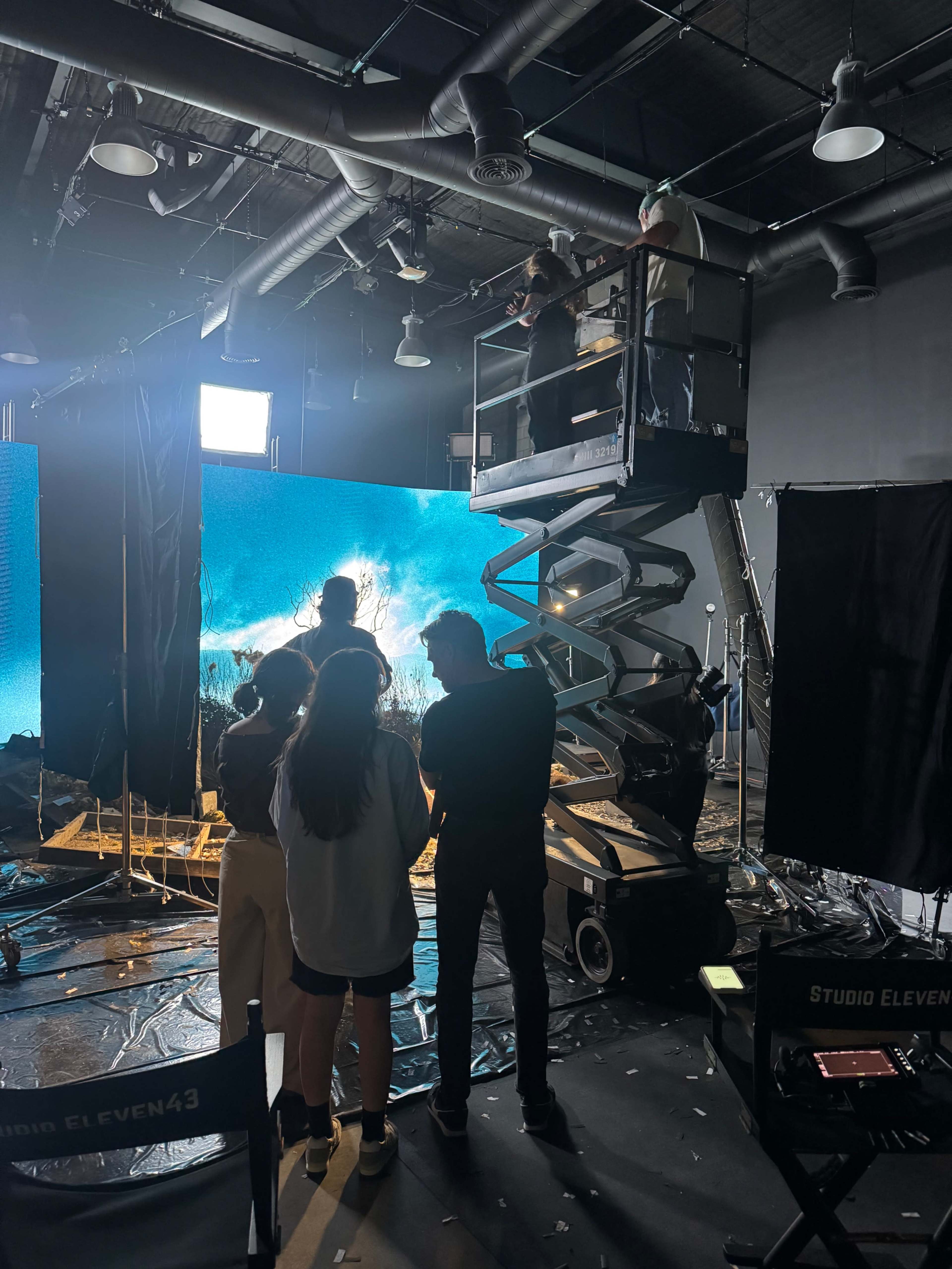 A film crew collaborates on a set featuring large digital screens, with one member elevated on a lift overseeing the scene.