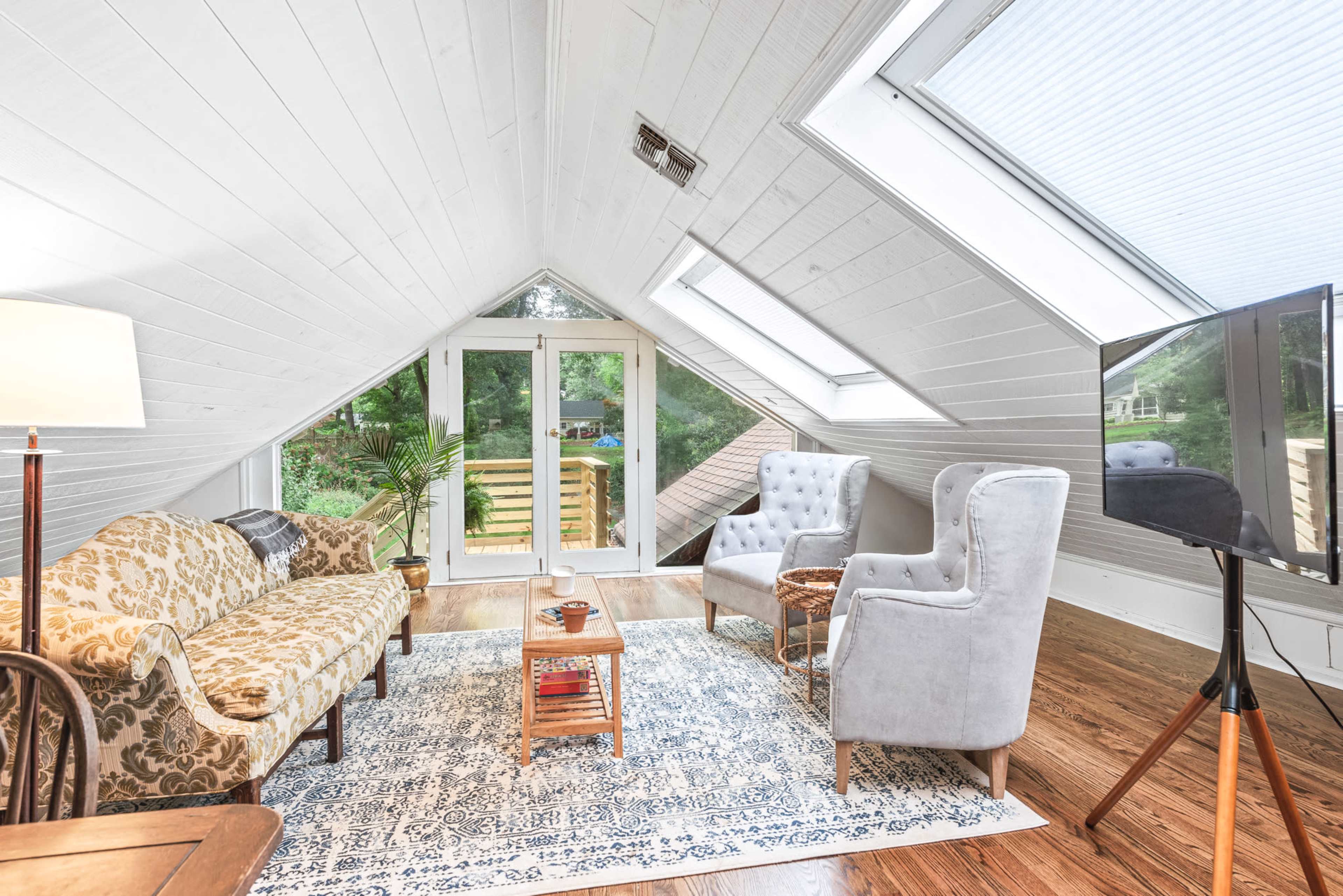 The image shows a cozy attic space furnished with a patterned sofa, two gray armchairs, a wooden coffee table, and large skylights illuminating the room.