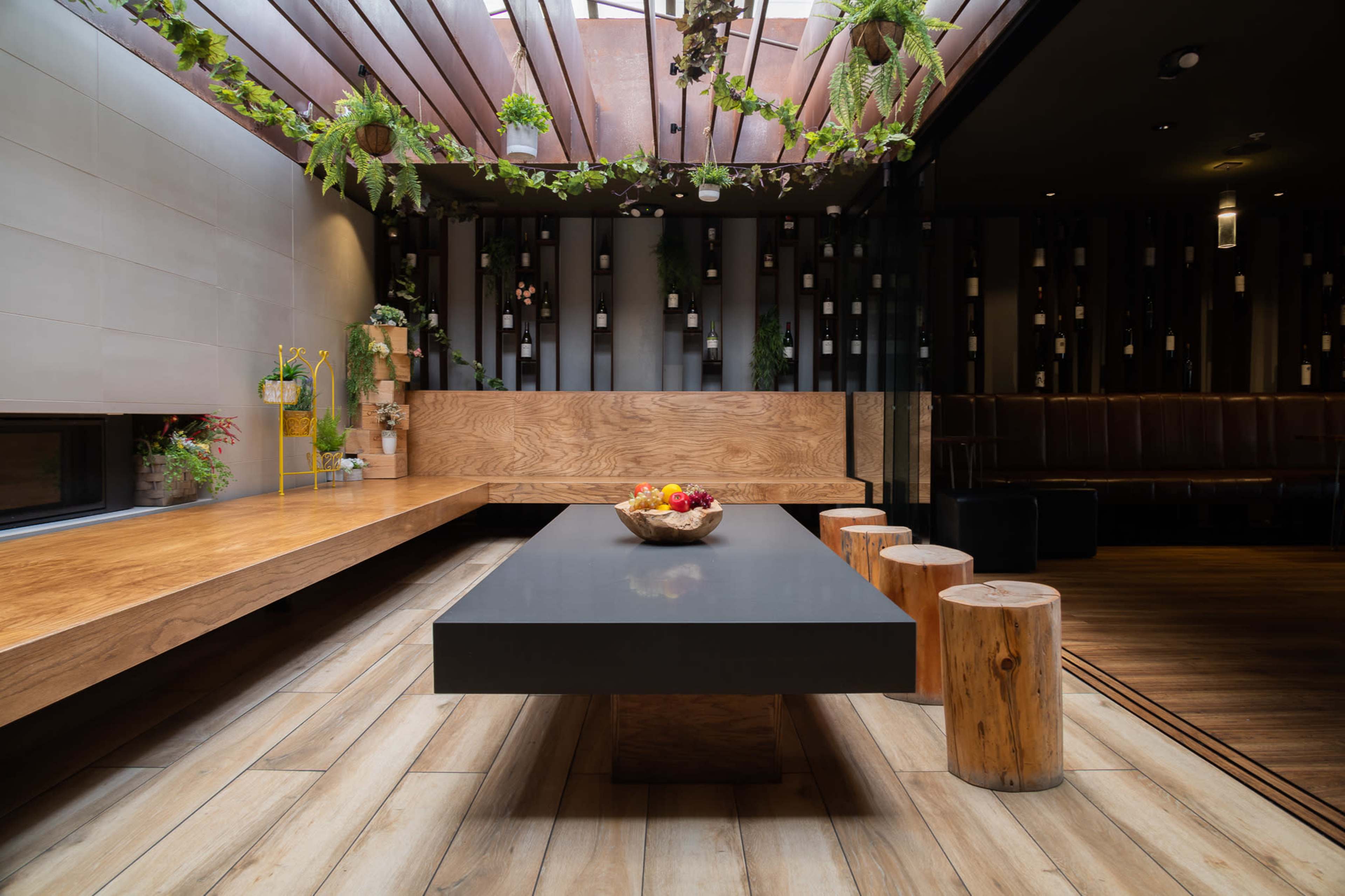 The image shows a modern interior space featuring a long black table surrounded by wooden stools and a bank of potted plants overhead.