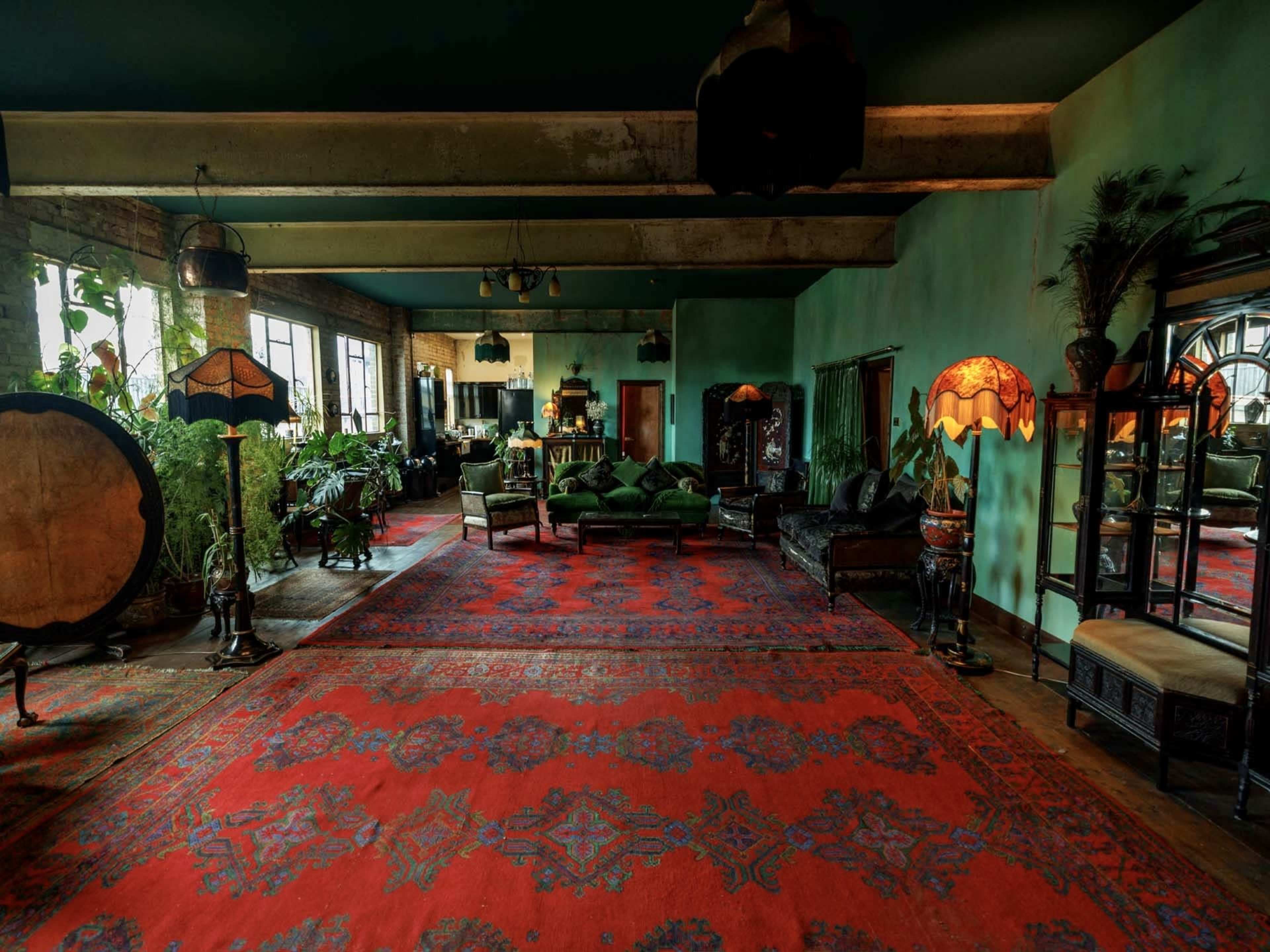 The spacious room features a large red area rug, multiple plants, vintage lamps, and a seating area with green upholstered furniture against a green-painted wall.