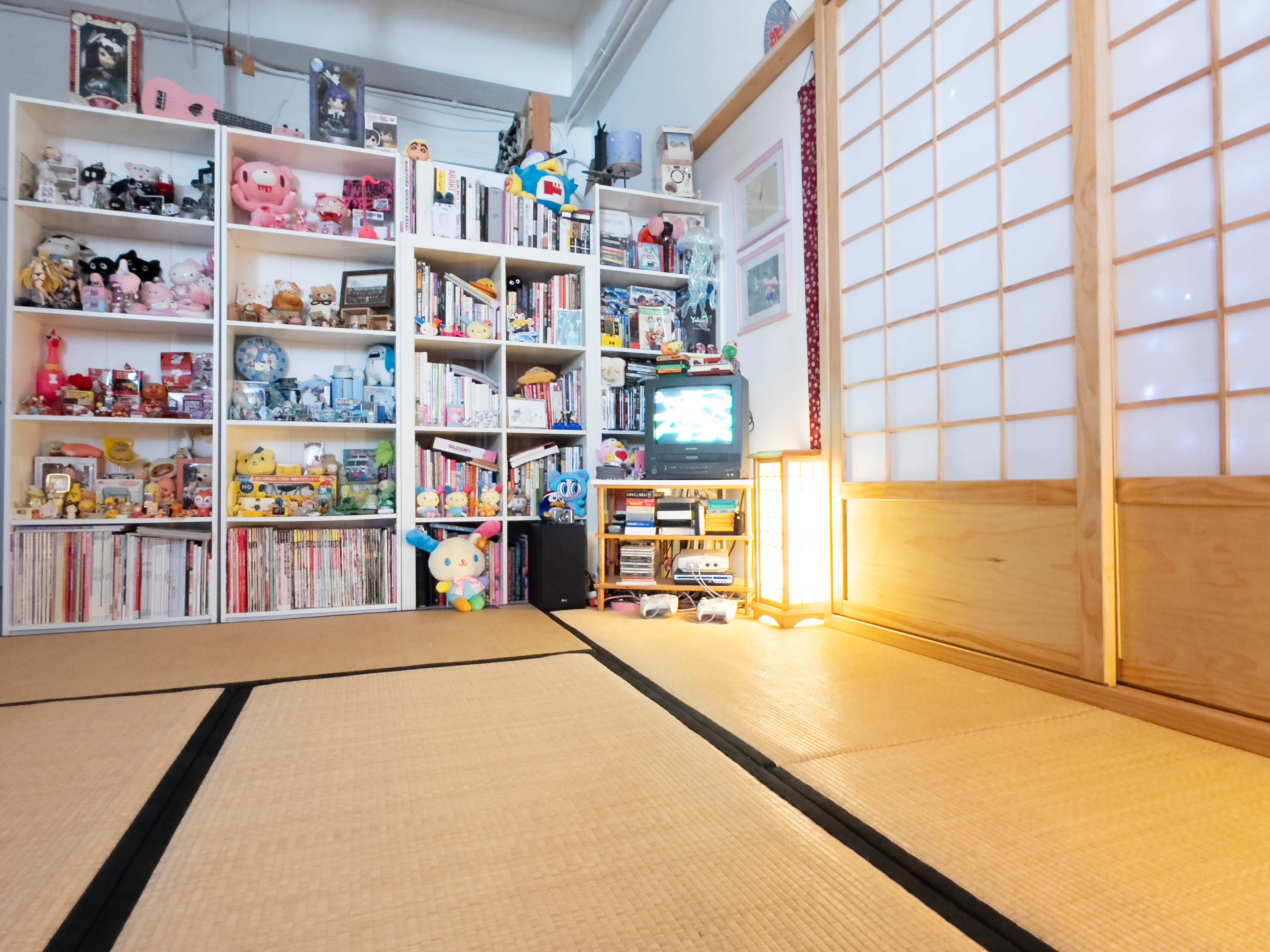 The image shows a cozy room with tatami flooring, featuring a wooden sliding door, a shelf filled with various collectibles, and a small TV beside a glowing lamp.