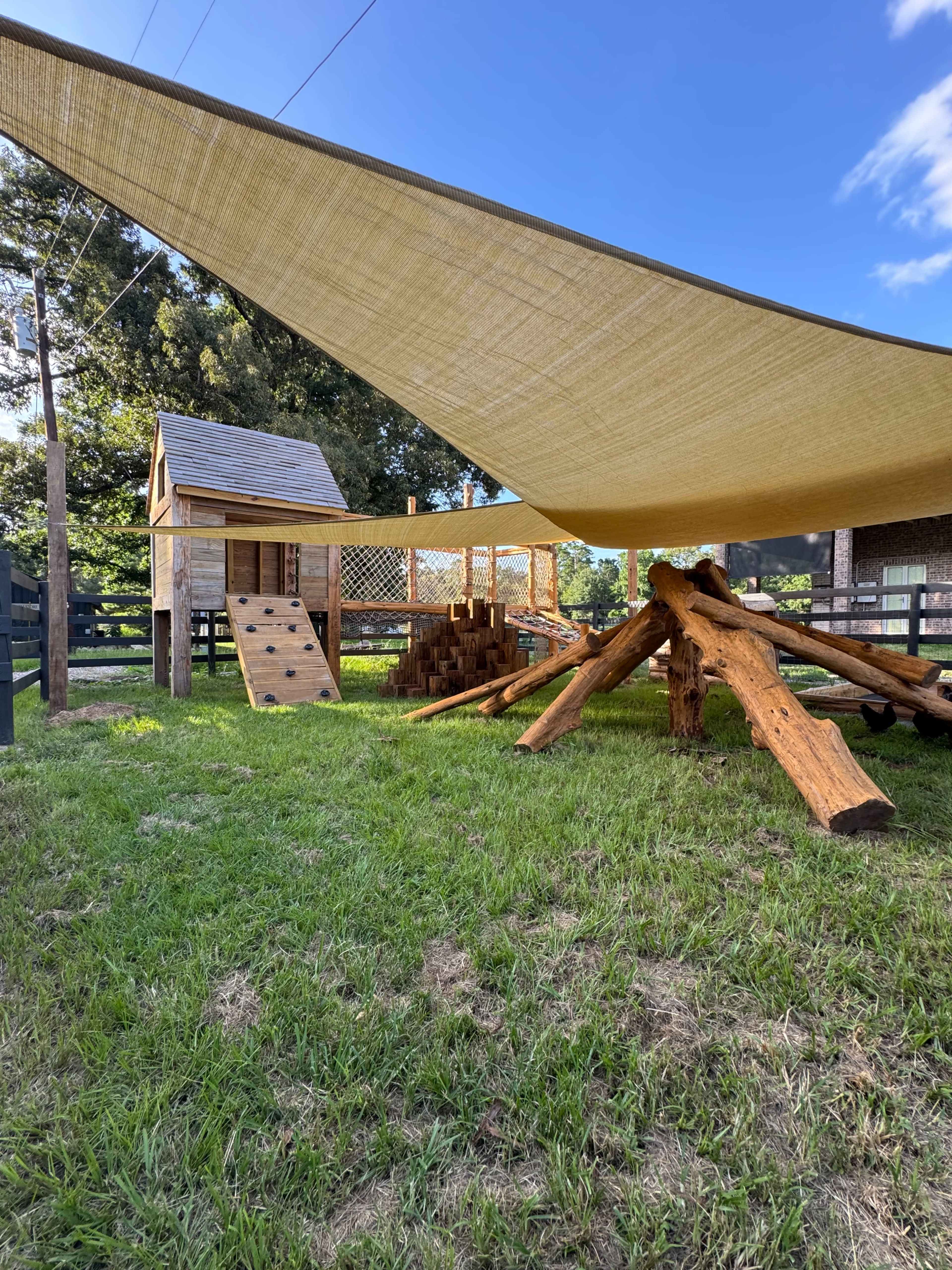 The image shows a wooden play structure with a slide and climbing wall under a large shaded canopy in a grassy area.