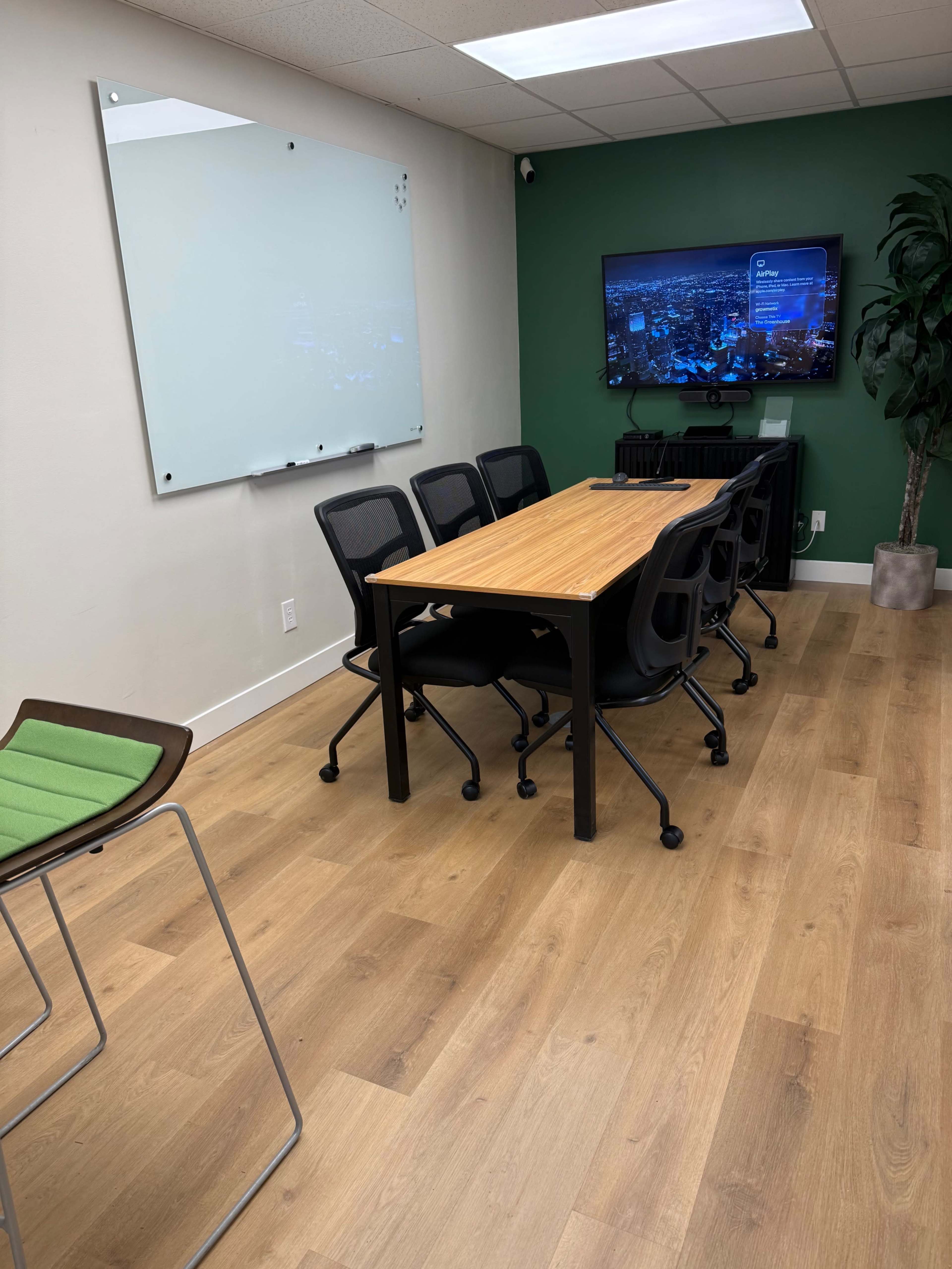 The image shows a small meeting room with a wooden table surrounded by black chairs, a green accent wall, a large screen displaying content, and a plant in the corner.