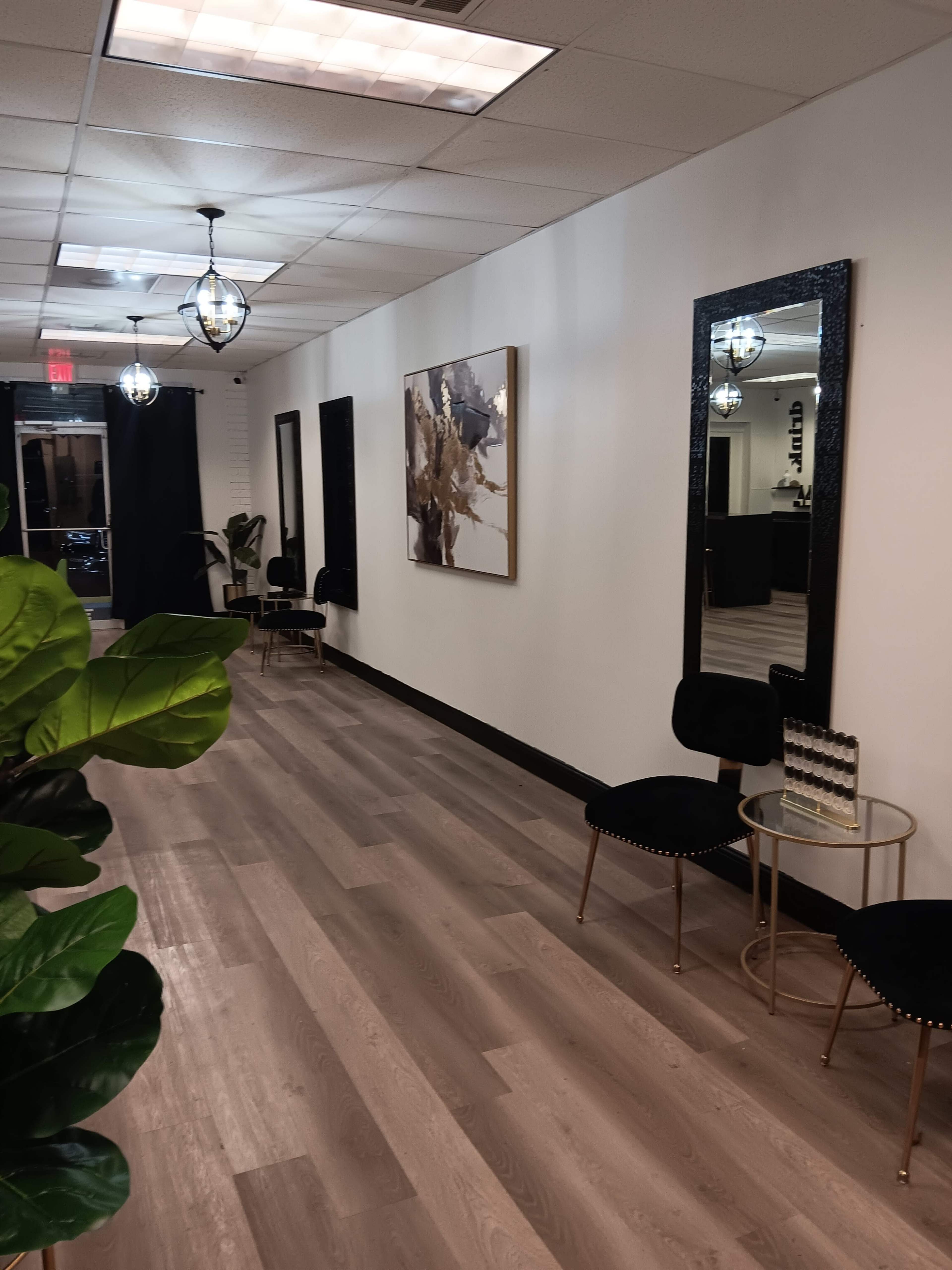 A well-lit hallway with a textured wall, featuring a large framed artwork, mirrors, and black chairs along the sides.