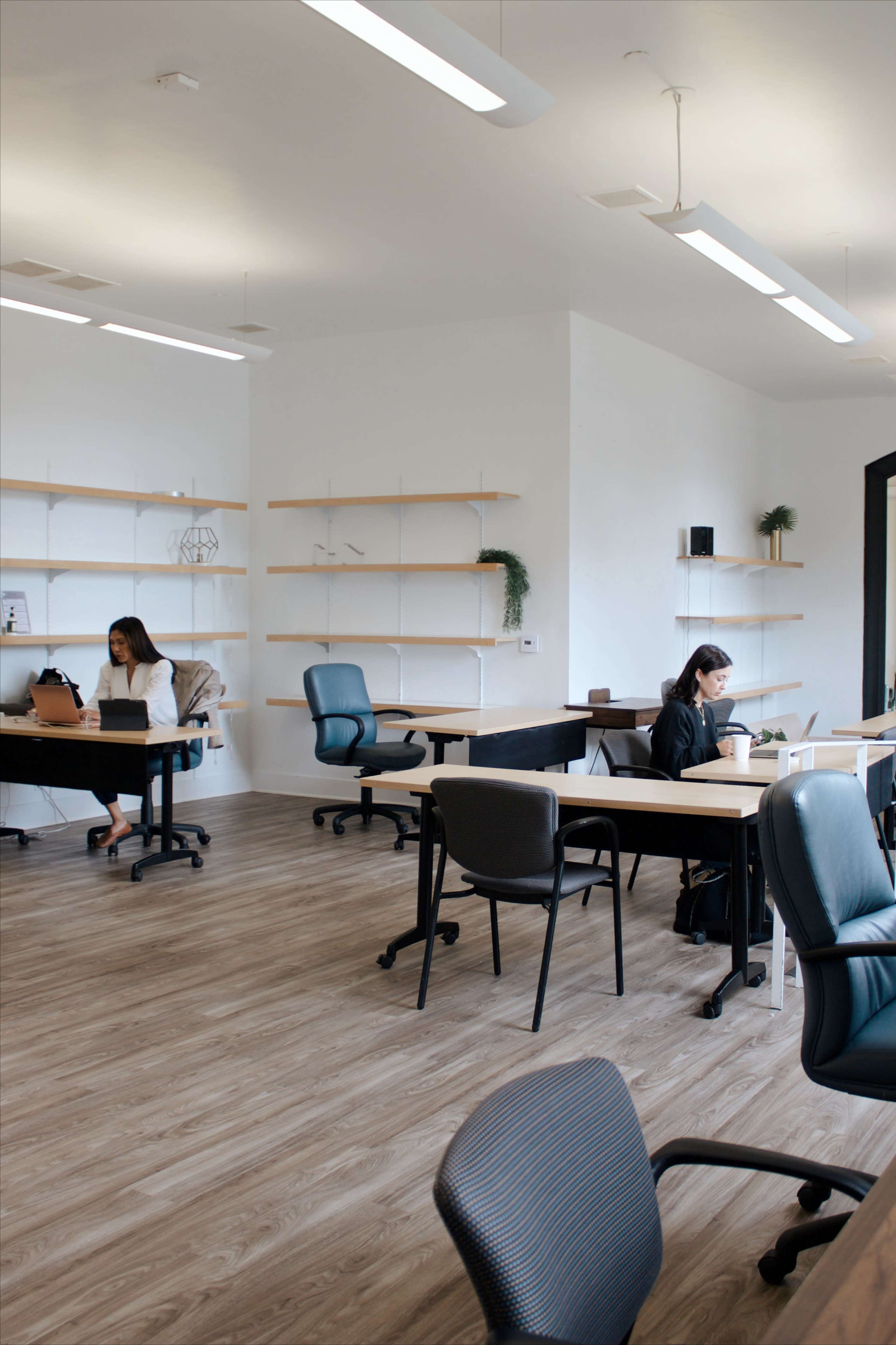 Two women work at separate desks in a modern office space with wooden floors and minimal shelving.