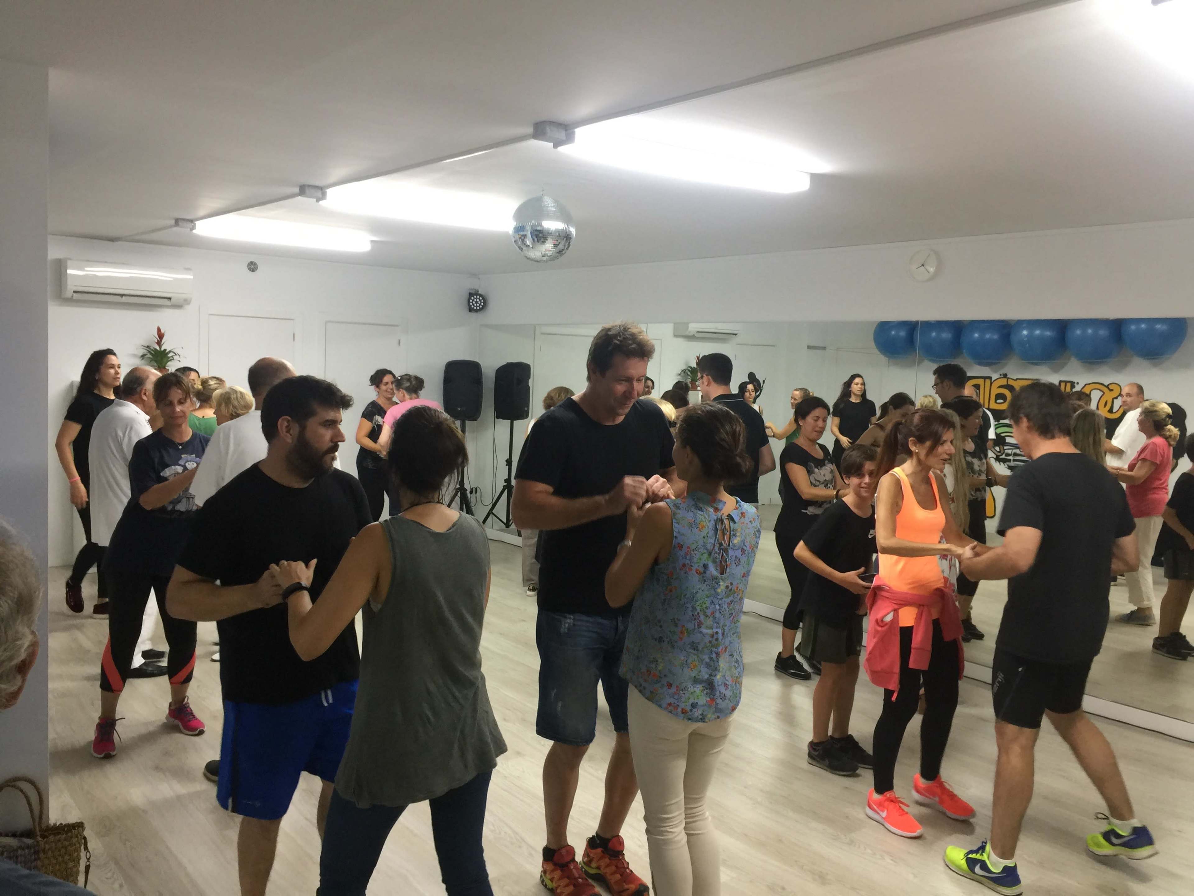 A group of people are dancing socially in a mirrored studio, with various participants engaged in partner dancing.