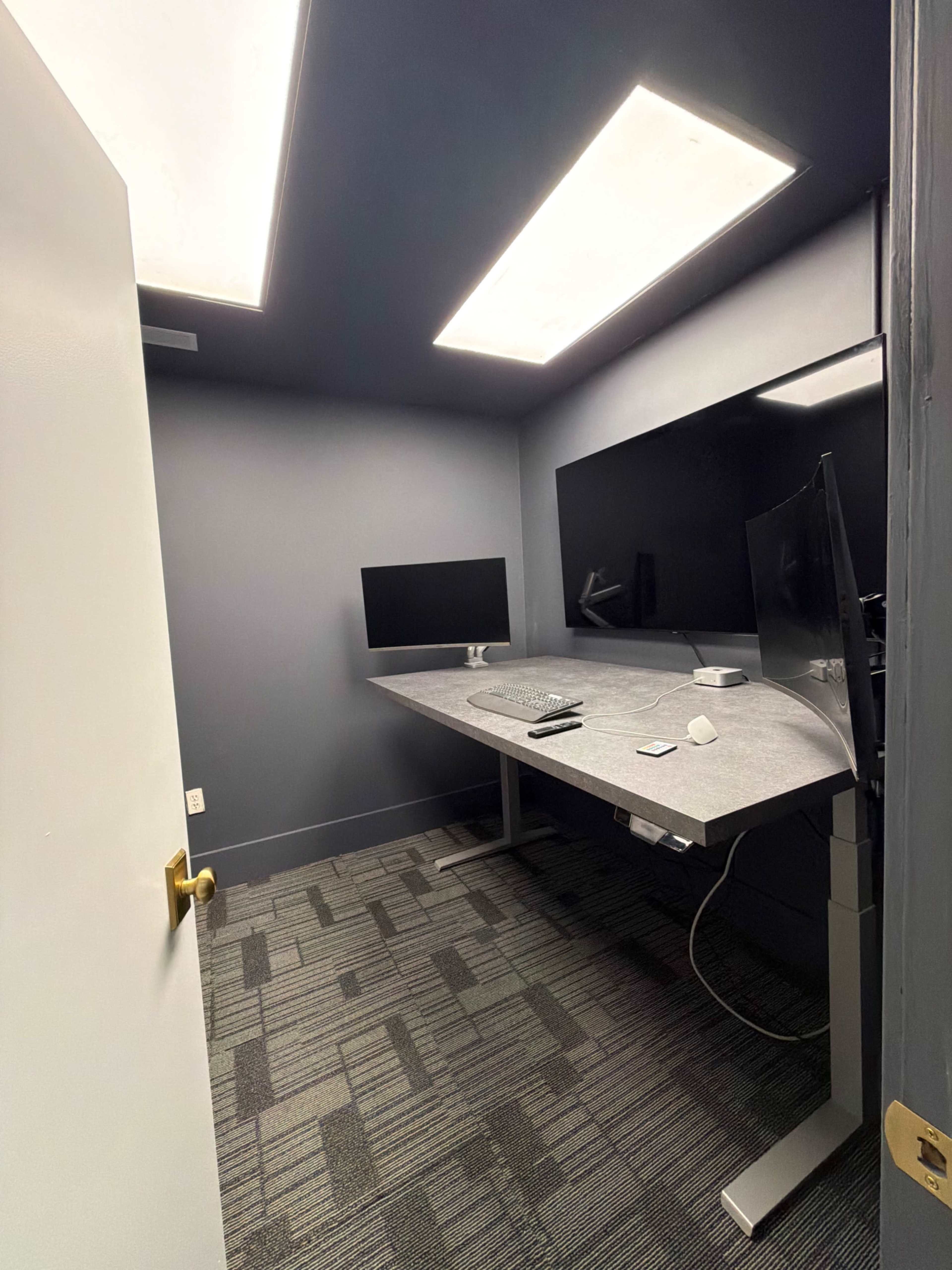 The image shows a small, modern office room with a large gray desk, two desktop monitors, and soft overhead lighting.