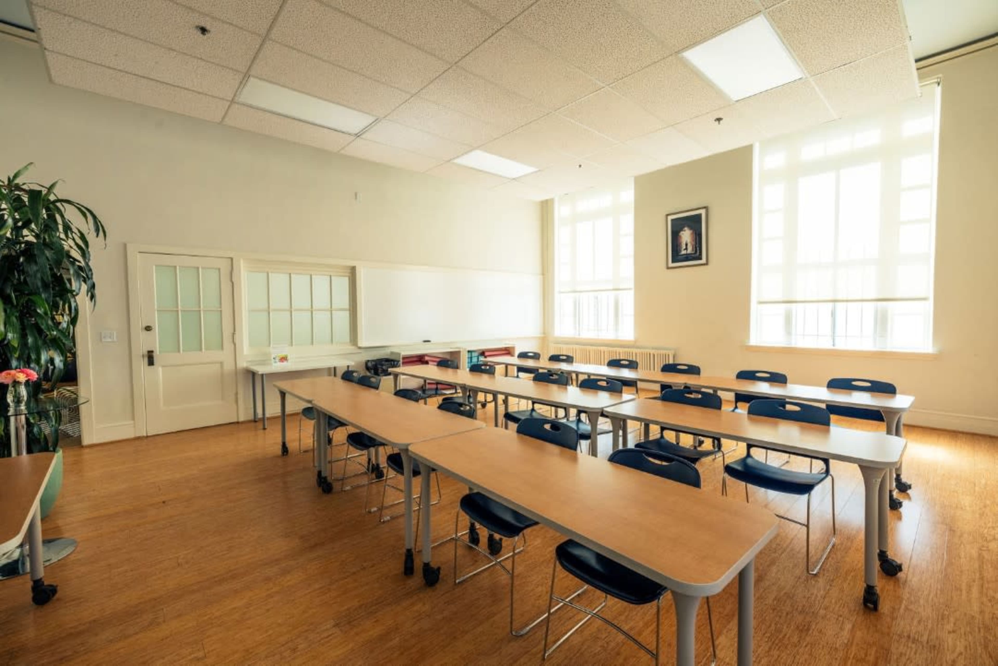 A classroom features several rows of tables and chairs, with large windows allowing natural light to enter.