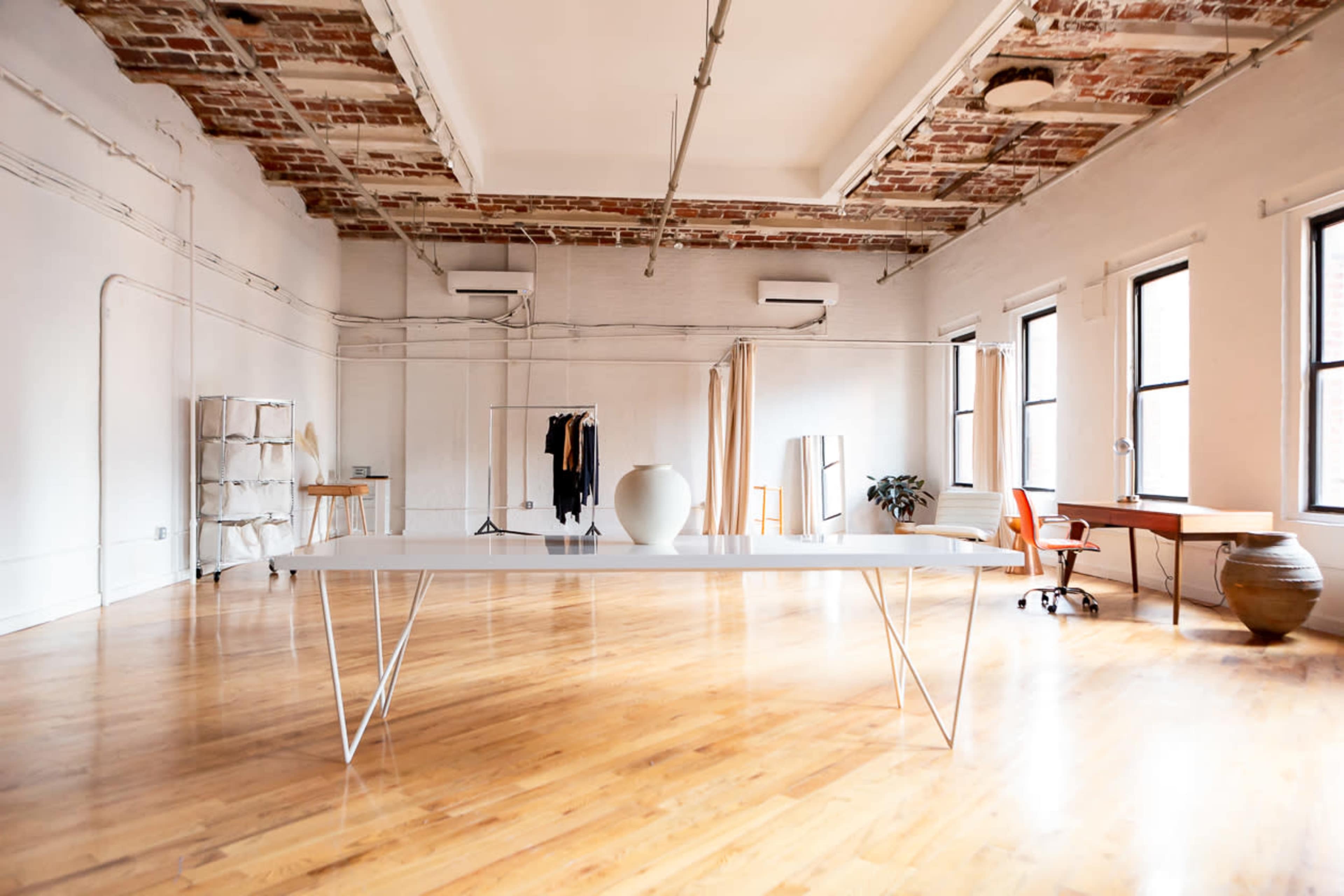 A spacious, well-lit interior with wooden floors, a long white table in the center, and various furnishings including a desk, chair, and clothing rack.
