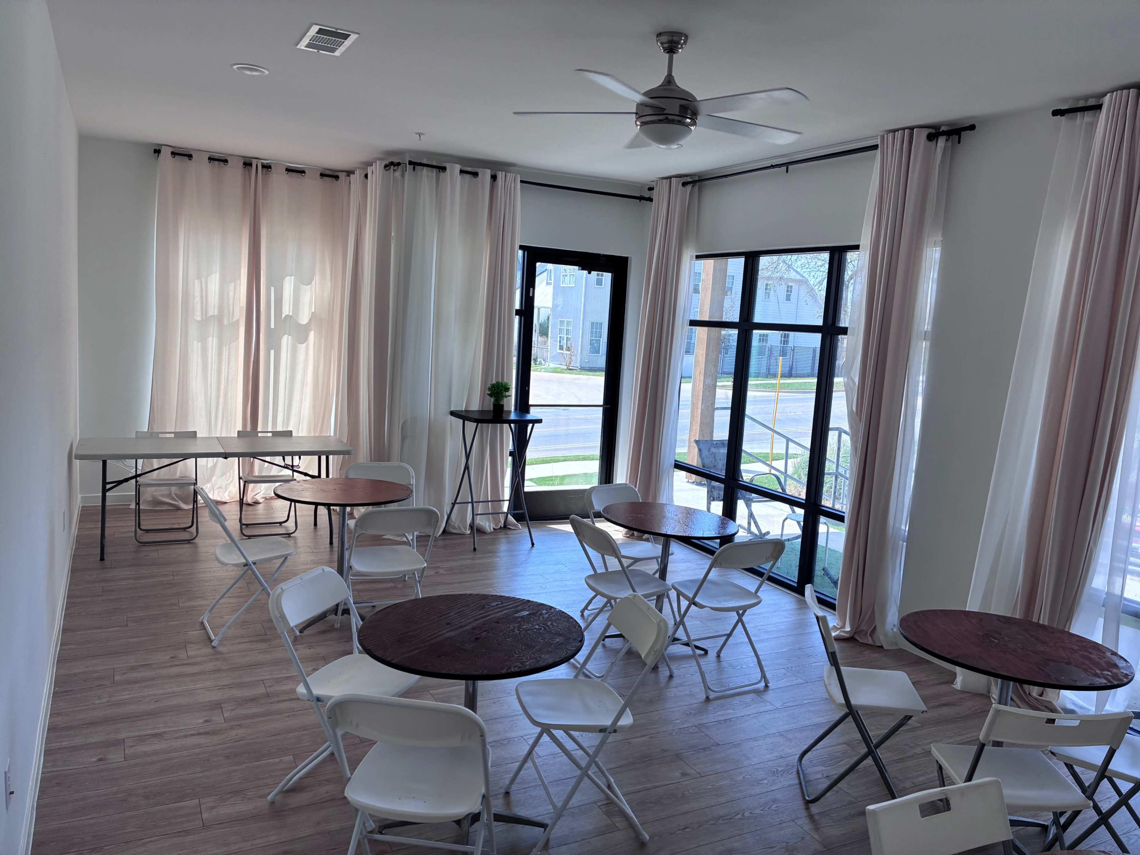 The image shows a bright, minimalistic room featuring several round tables with chairs, large windows dressed with sheer curtains, and a small indoor plant on a tall table.