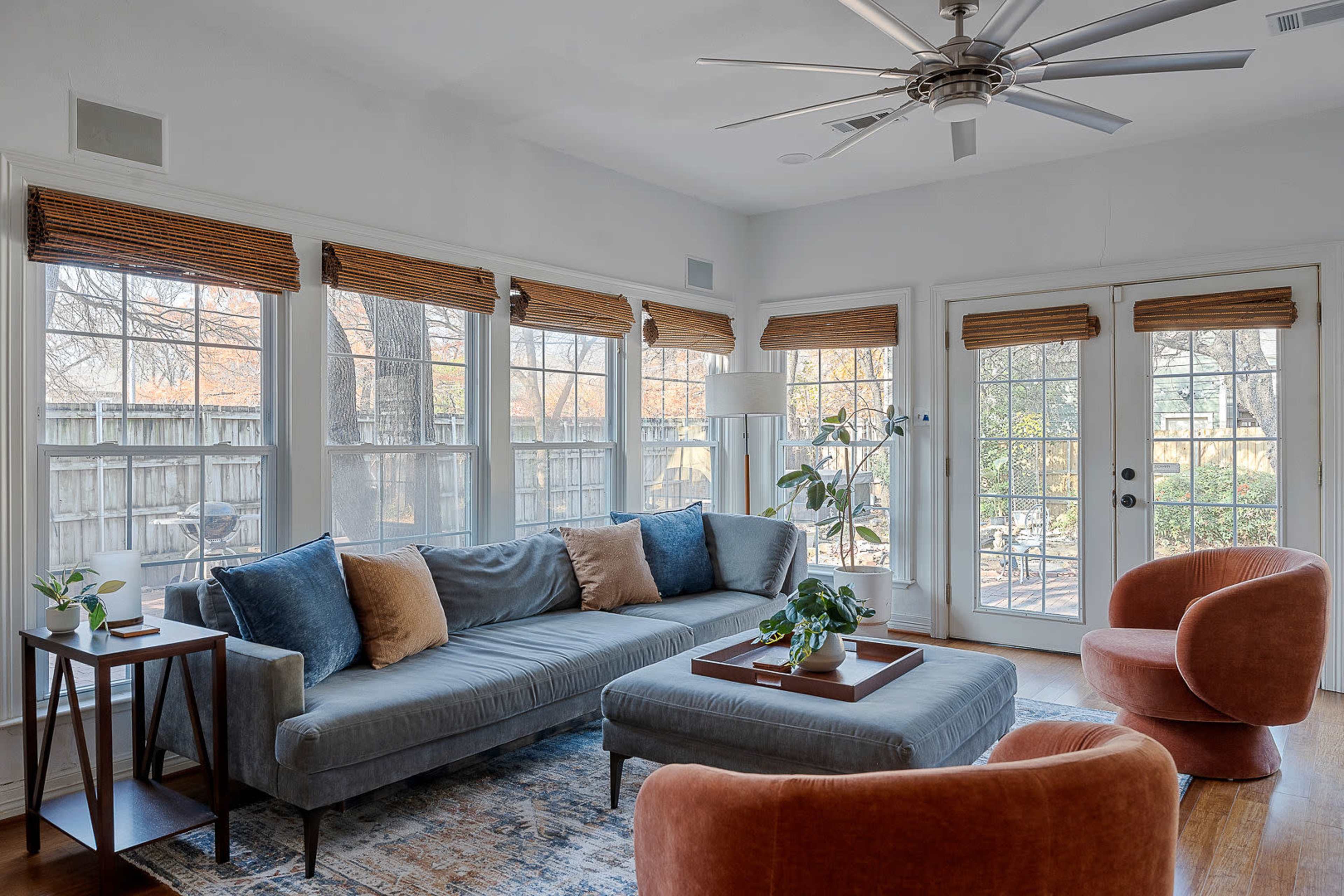The living room features a gray sectional sofa, orange accent chairs, large windows with wooden blinds, and a coffee table in the center.