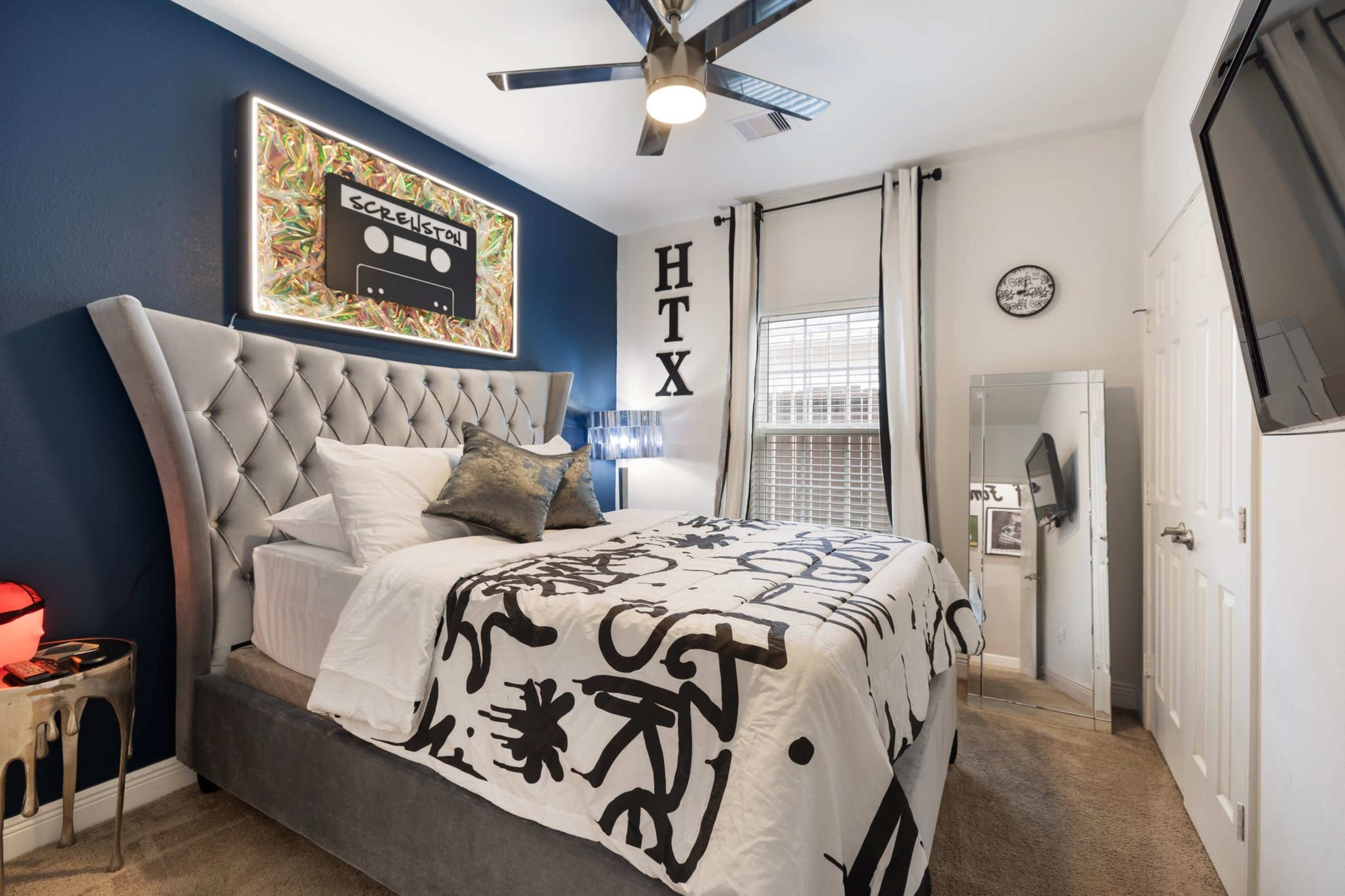 The image shows a neatly arranged bedroom featuring a tufted bed with a decorative black and white bedspread, a colorful artwork on the wall, and modern furnishings including a mirror and a wall-mounted TV.