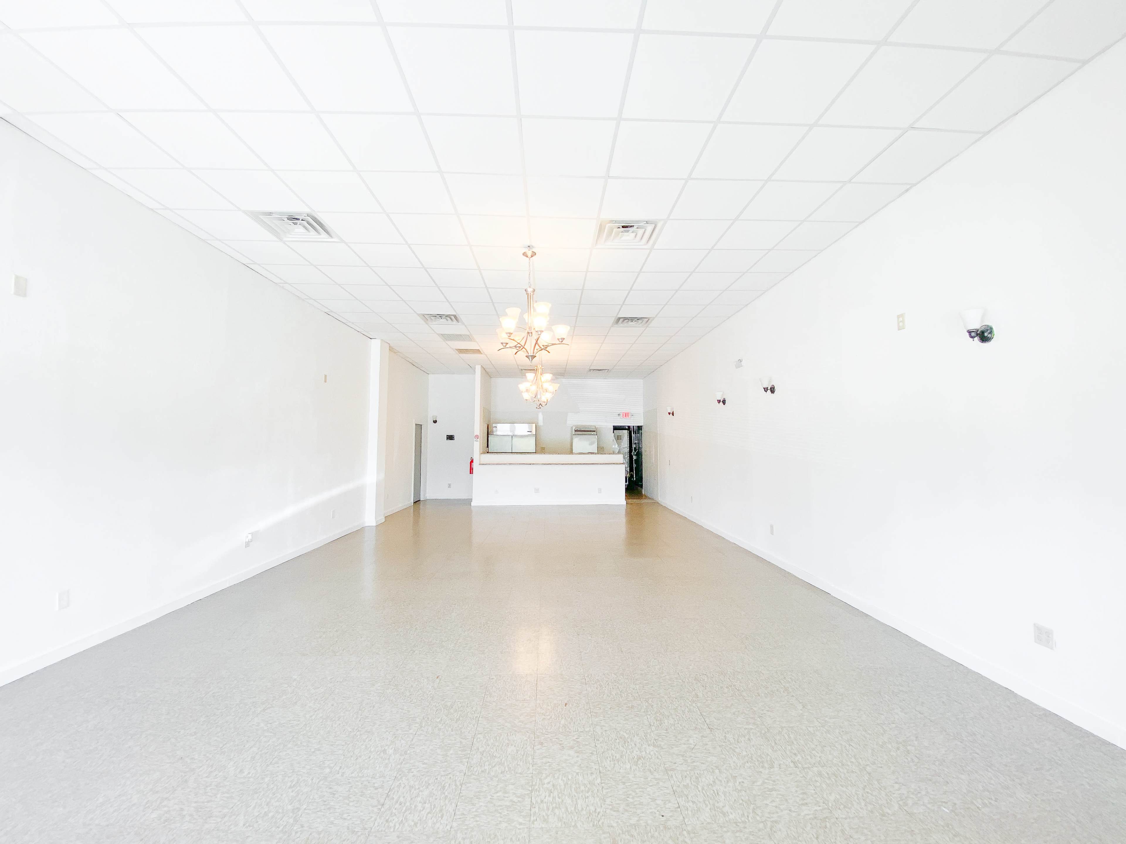 The image shows a spacious, empty room with light-colored walls, a chandelier, and a counter at the back.