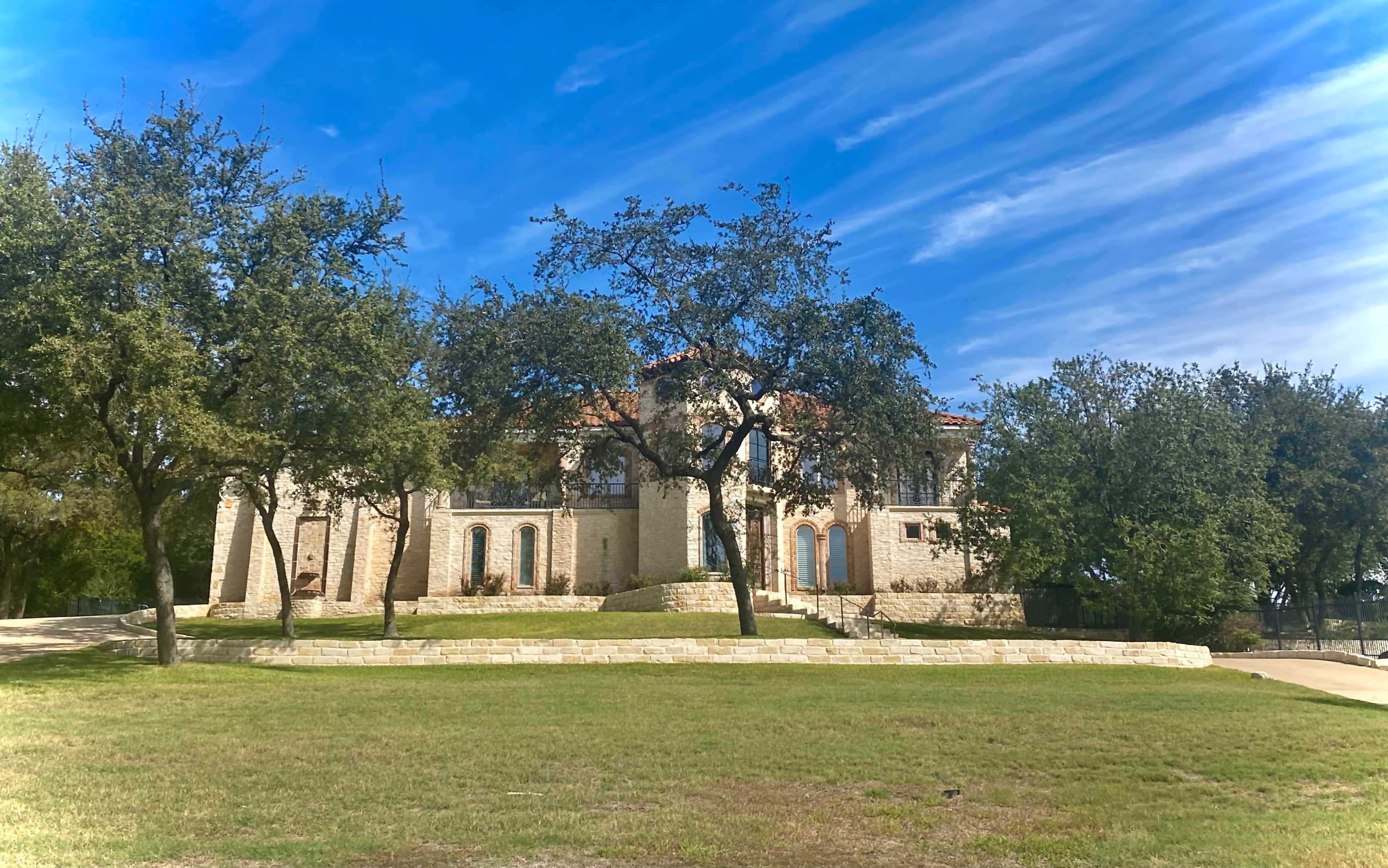 A large stone mansion with a red tile roof is set against a backdrop of blue sky and trees on a grassy hillside.