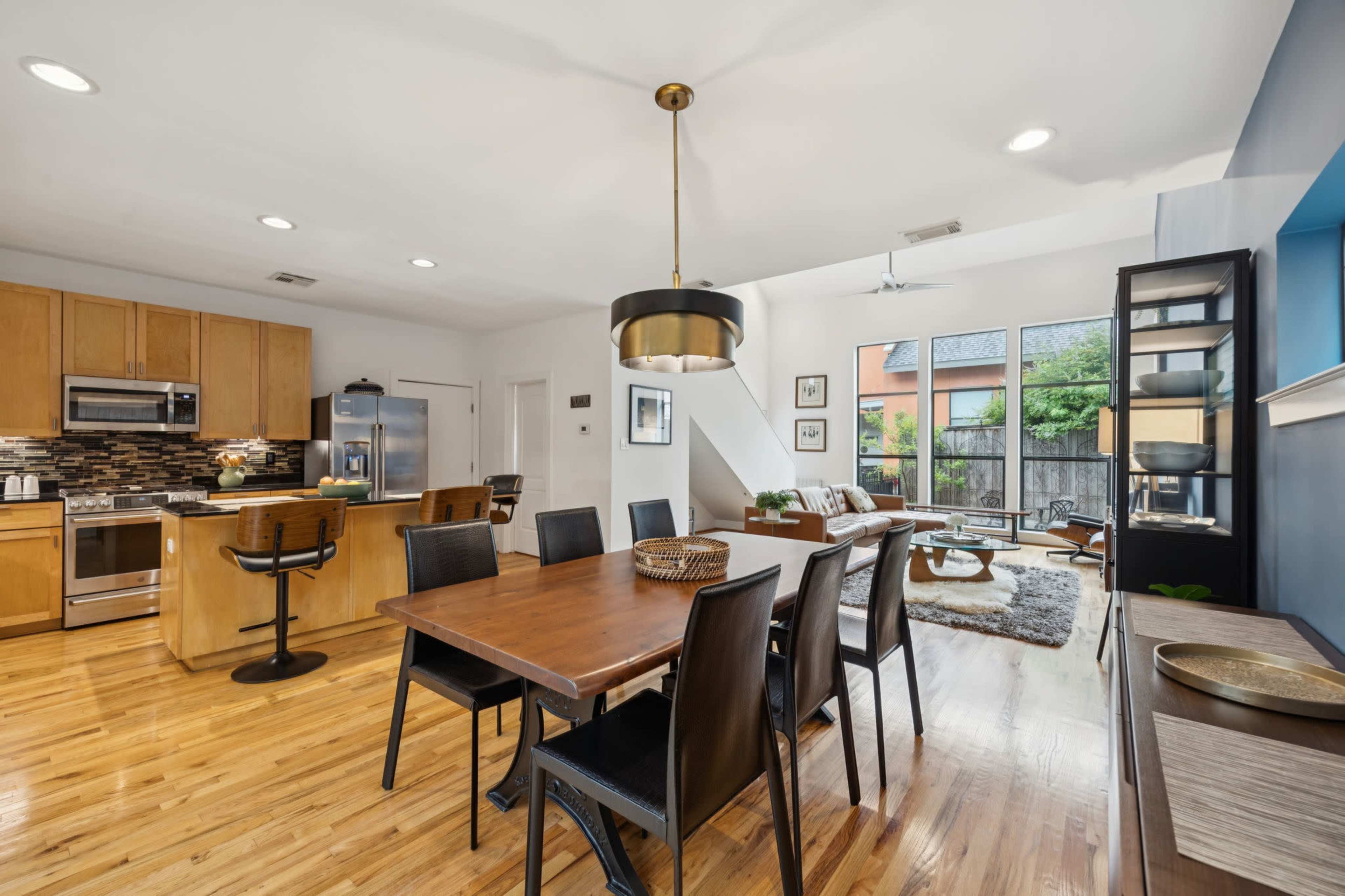 The image shows a modern kitchen and dining area featuring wooden cabinets, a large dining table, and a living space with a view of a backyard.
