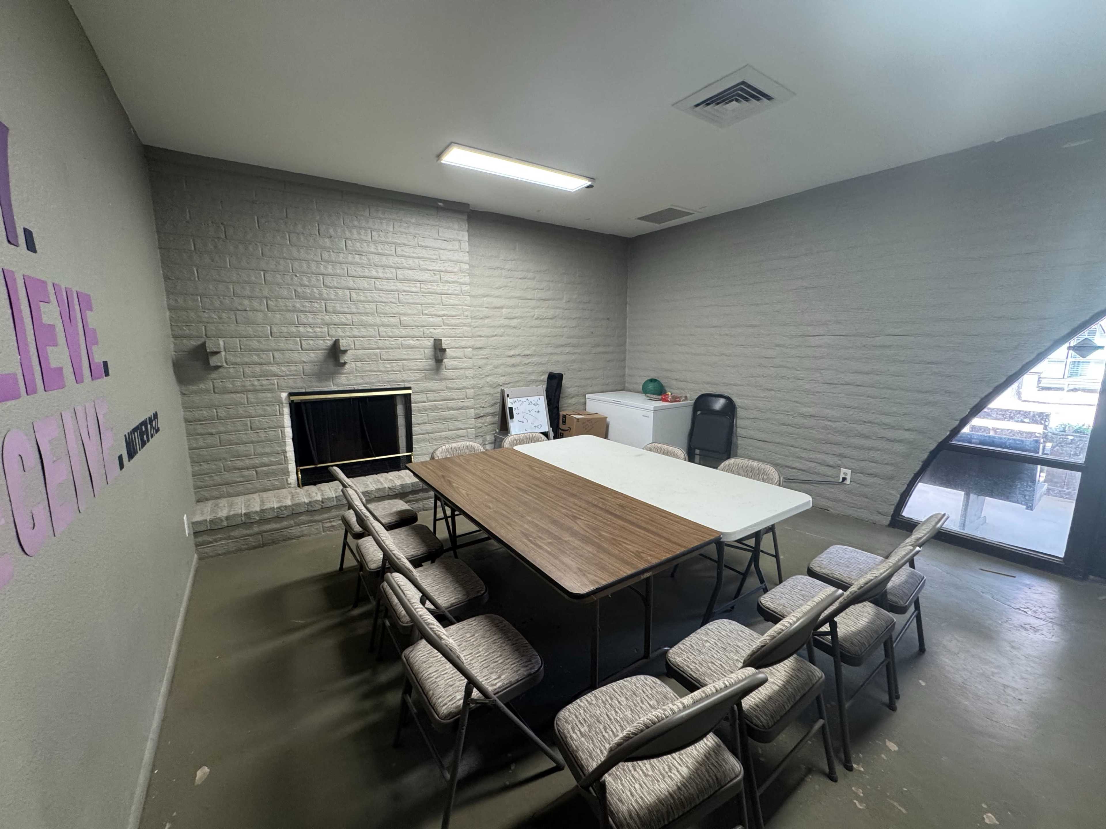 A rectangular table surrounded by eight chairs sits in a sparsely furnished meeting room with a fireplace and a door leading outside.
