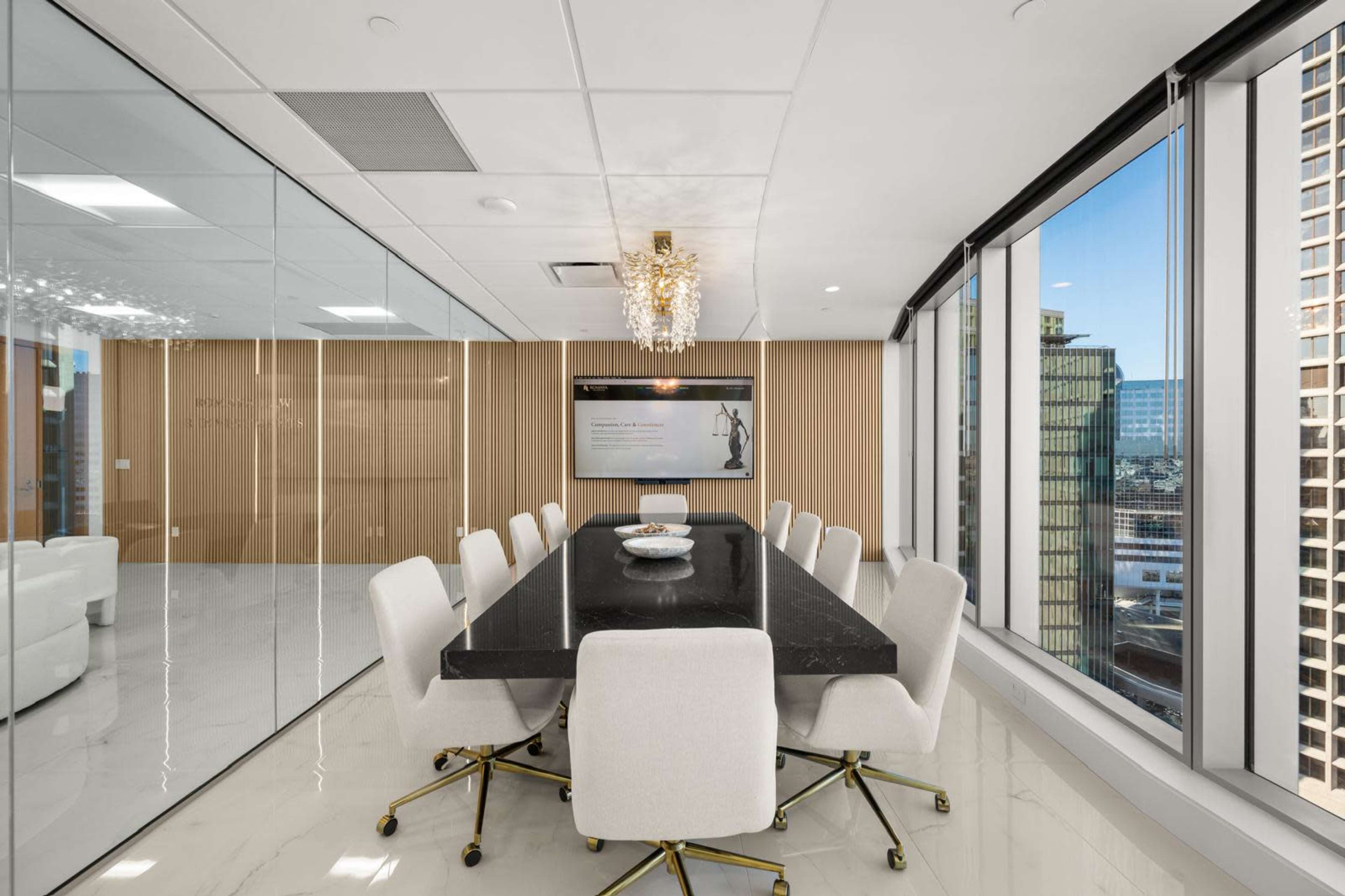 The image shows a modern conference room with a long black table surrounded by white chairs, large windows offering a city view, and an elegant chandelier overhead.