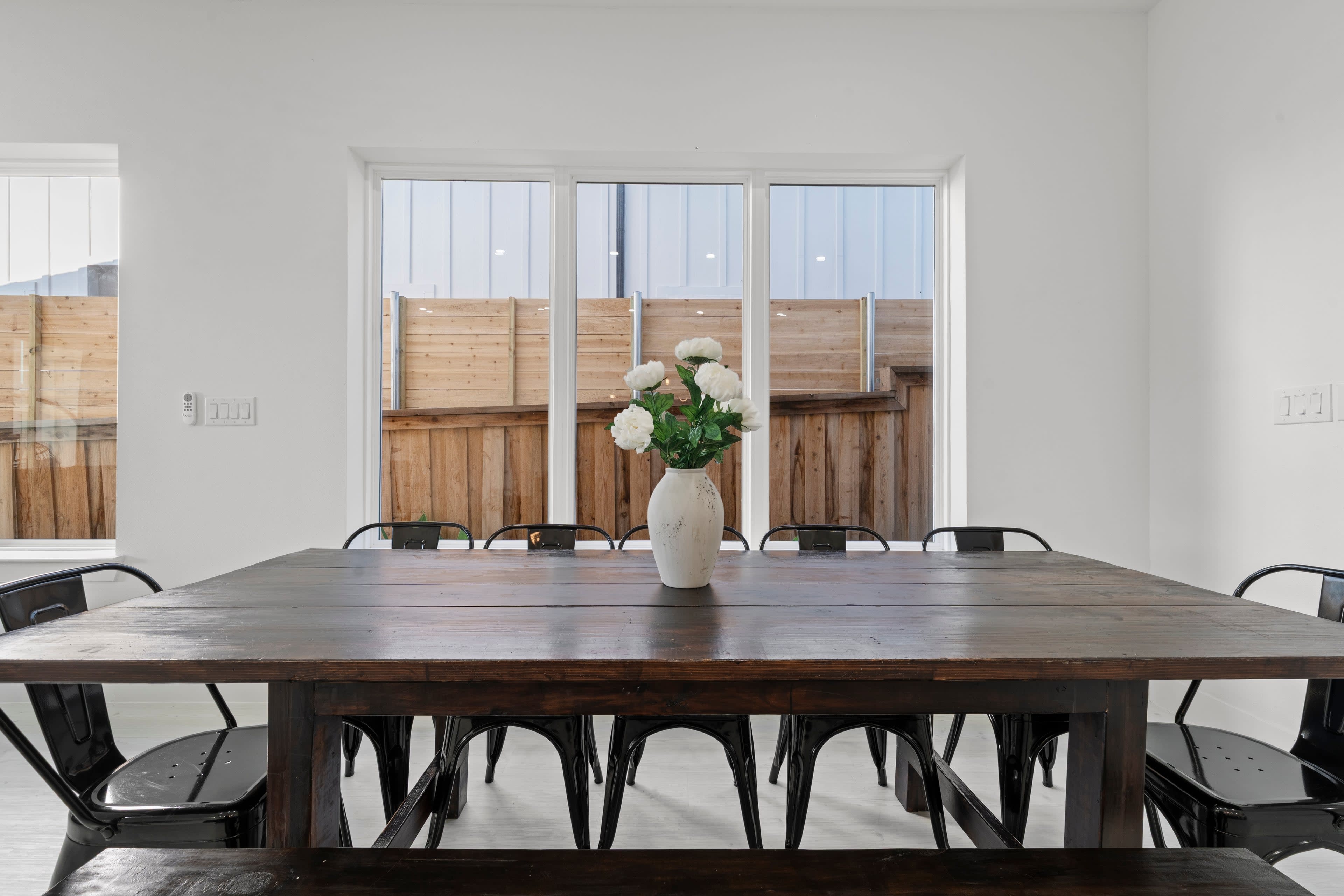 A wooden dining table with a vase of flowers is set in a bright room facing a fenced outdoor area through large windows.