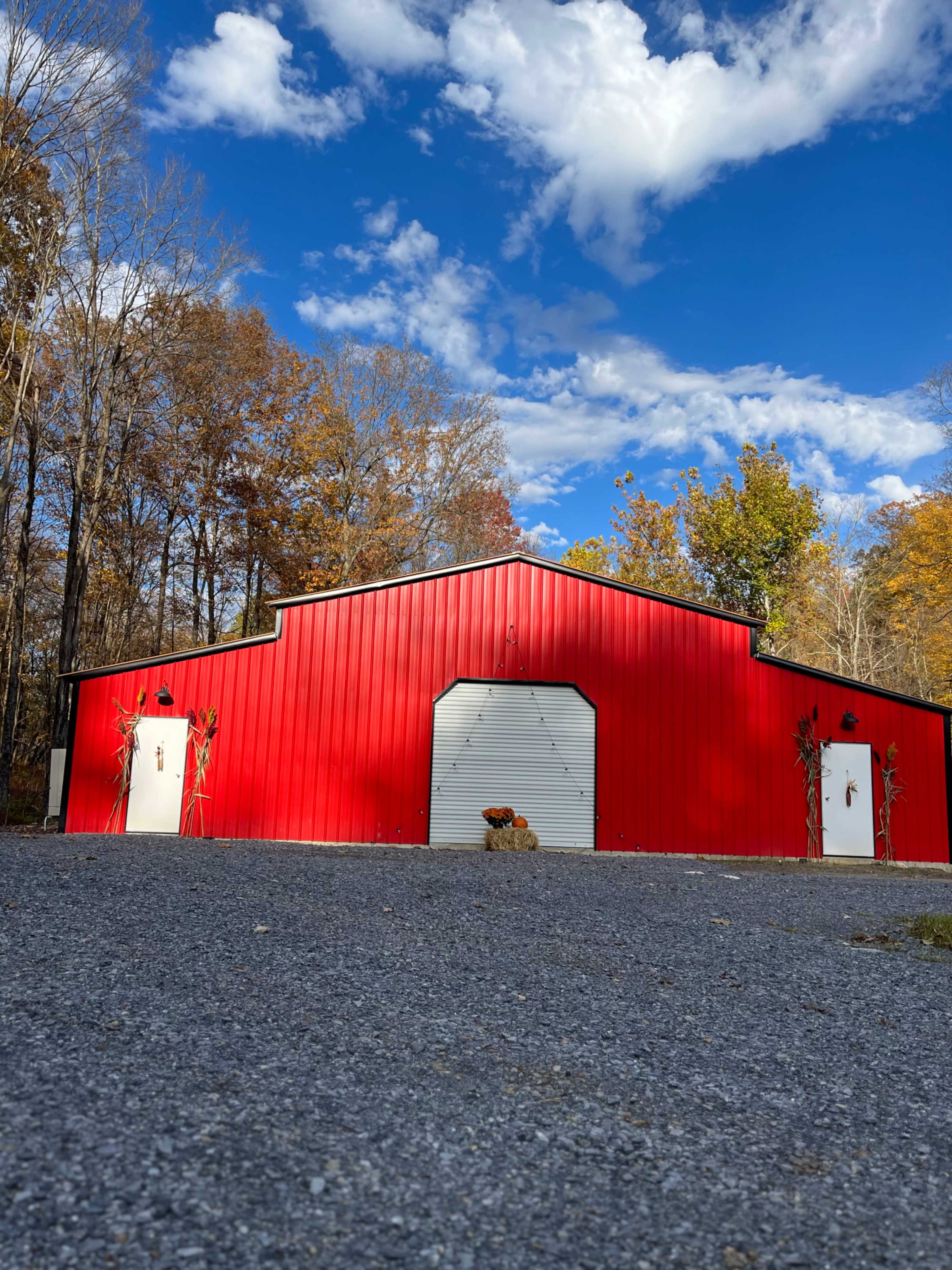 A large red barn with a gray gravel driveway is surrounded by trees with autumn foliage under a partly cloudy sky.