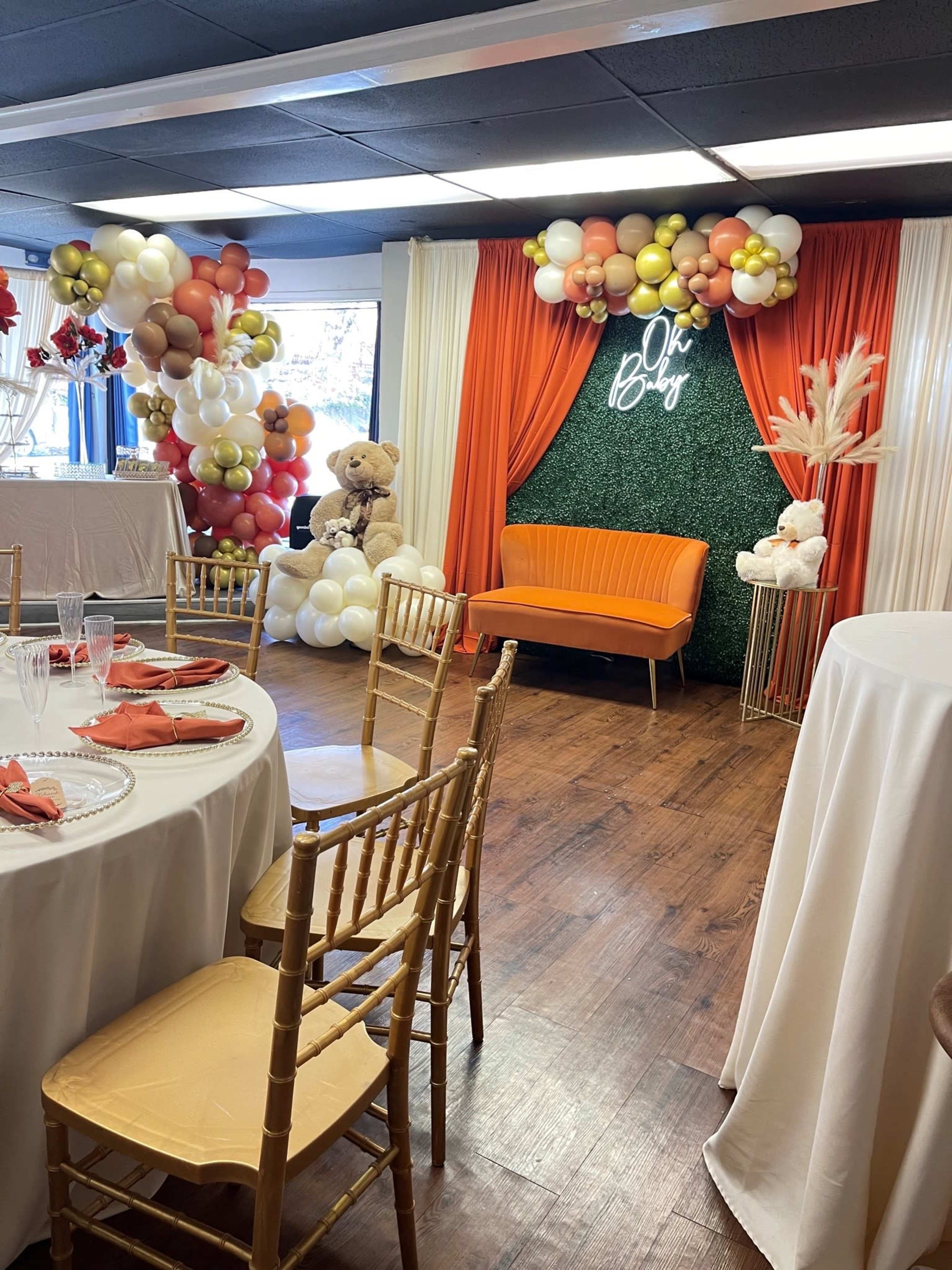 A decorated event space features an orange sofa against a greenery backdrop, surrounded by balloon arrangements and a table set for a celebration.