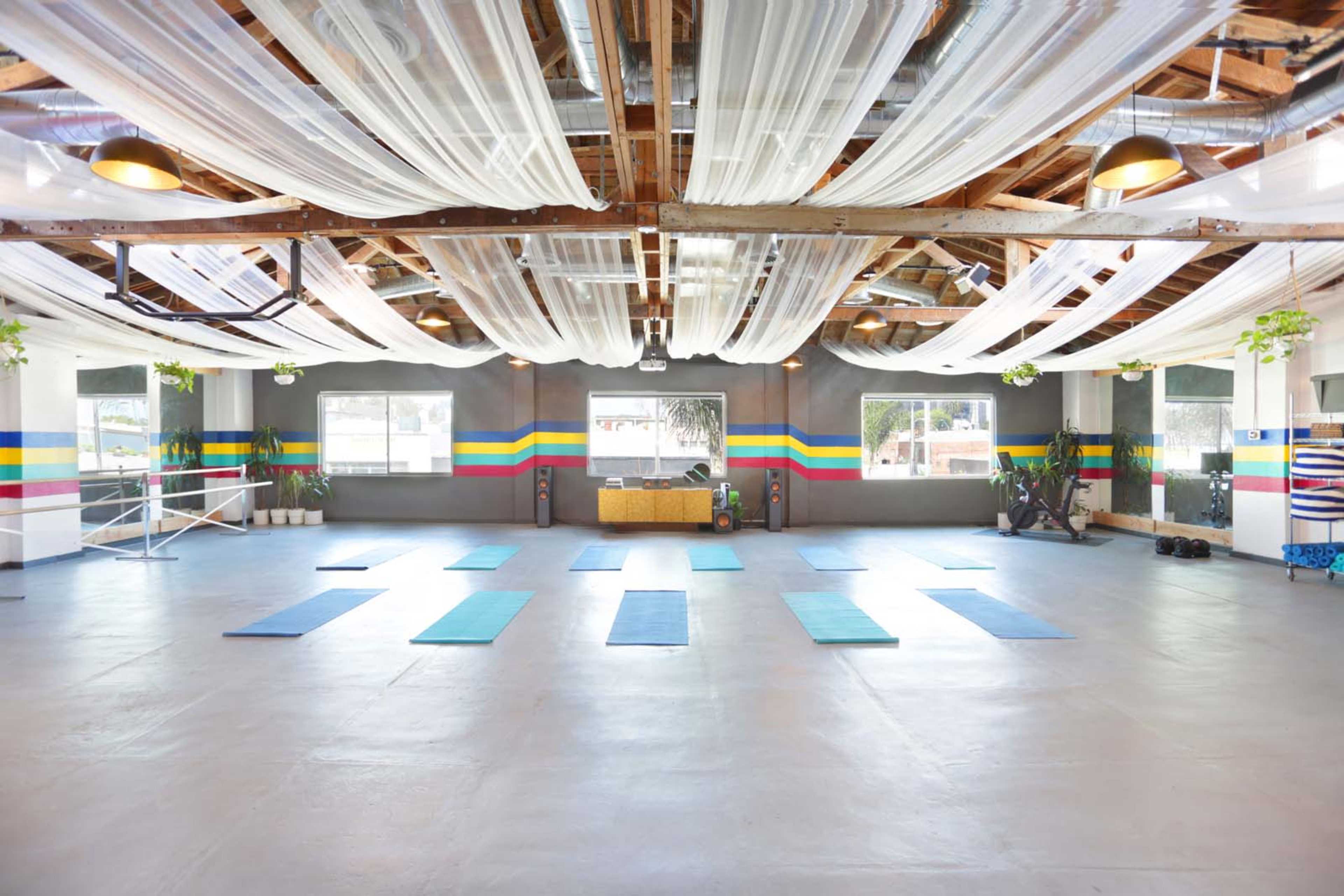 The image shows a spacious indoor area designed for yoga or fitness, featuring blue mats arranged on the floor, colorful stripes on the walls, and fabric draped from the ceiling.