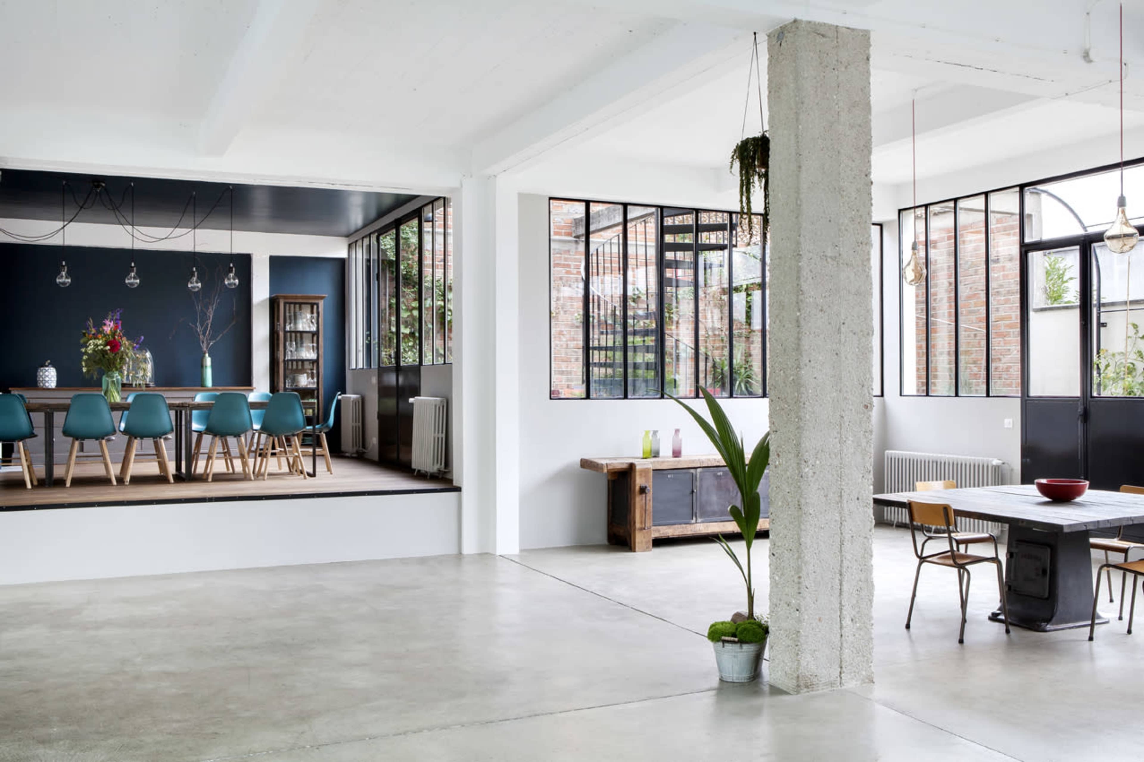 A spacious interior with a modern dining area featuring a large table and blue chairs, and an adjacent room with large windows and plants, separated by a concrete column.