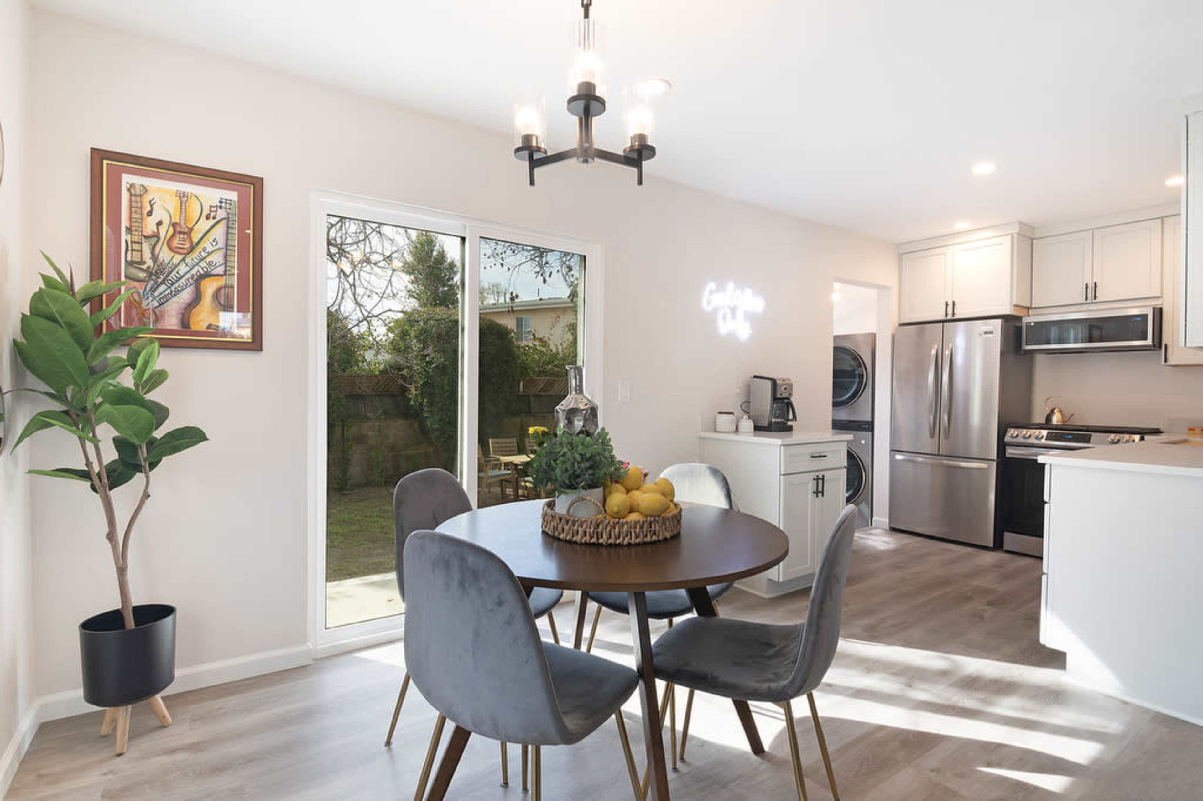 A dining area features a round table with gray chairs, a large window overlooking a garden, and modern kitchen appliances in the background.