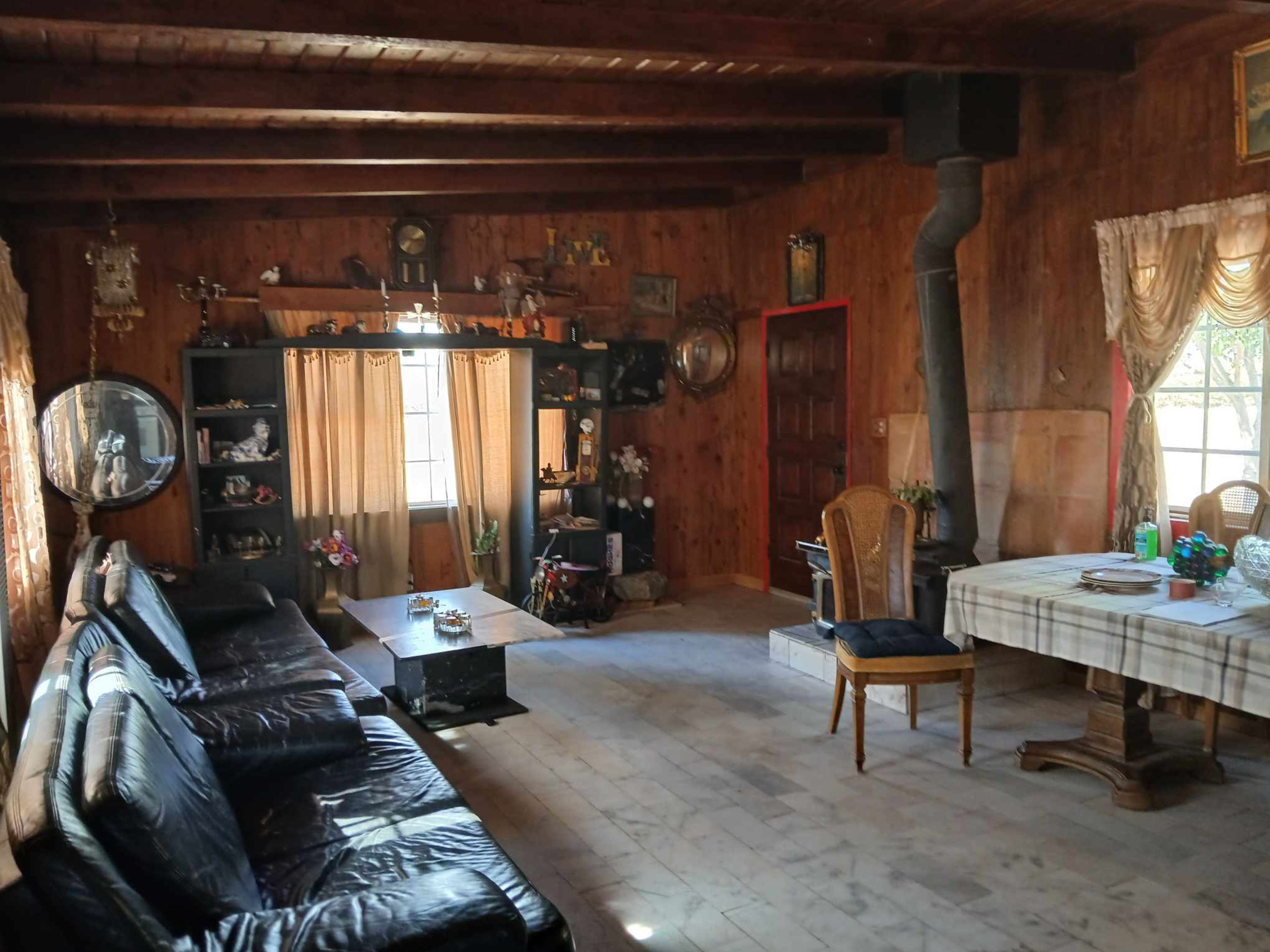 The image shows a rustic wooden living room featuring a black leather sofa, a wooden coffee table, a dining area with a table and chairs, and various decorations on the walls and shelves.