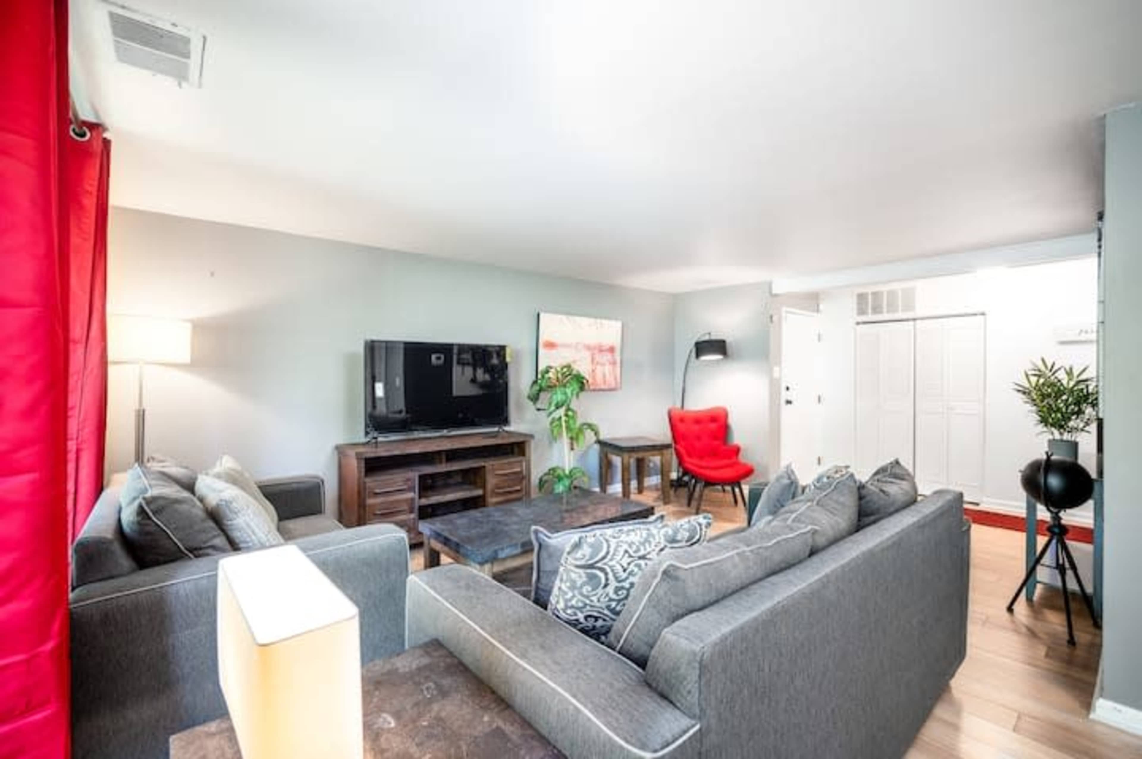 The living room features a large gray sectional sofa, a red accent chair, a television on a wooden stand, and a potted plant, with light hardwood flooring and bright red curtains.