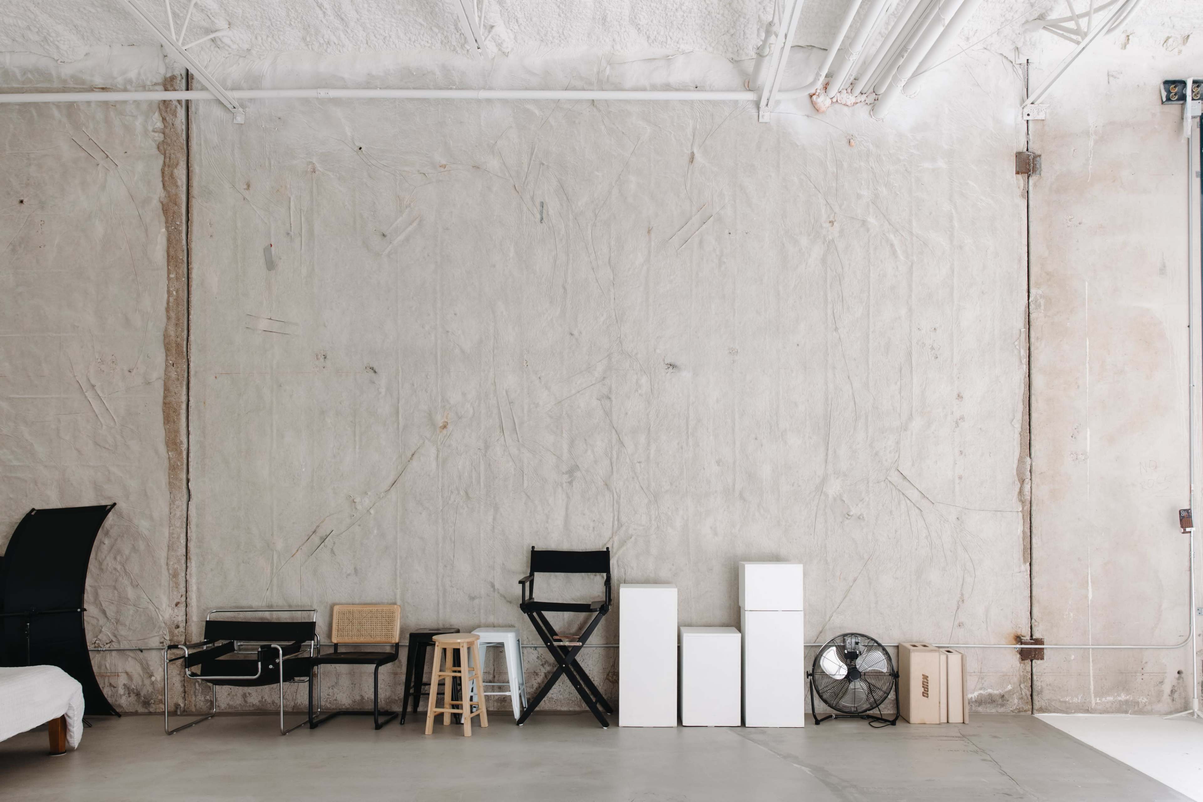 The image shows a minimalistic interior with an unfinished concrete wall and a row of various furniture pieces, including chairs, boxes, and a fan, arranged along the floor.
