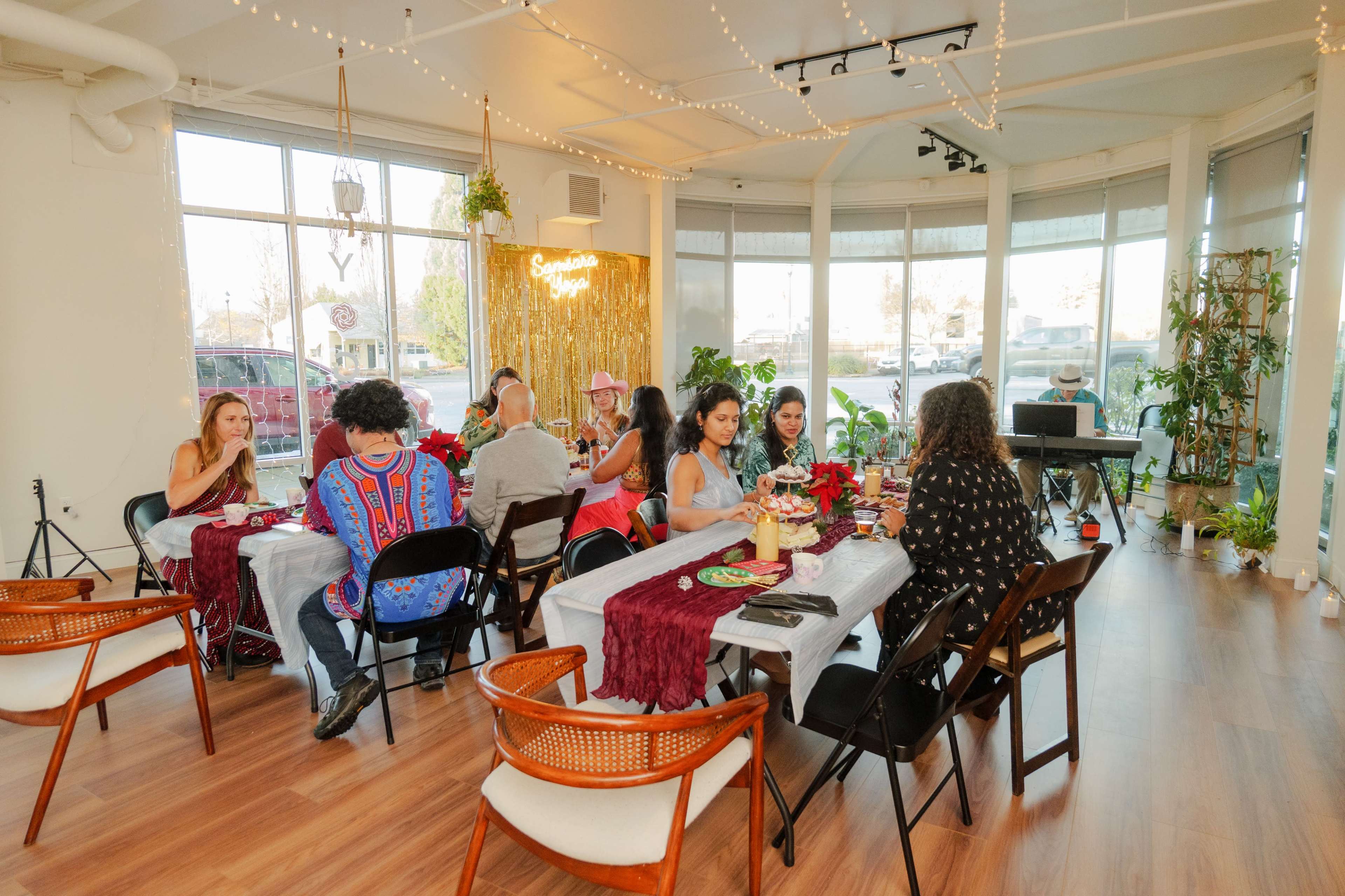 A group of people are seated around several tables in a warmly lit dining space, decorated with plants and festive decor.