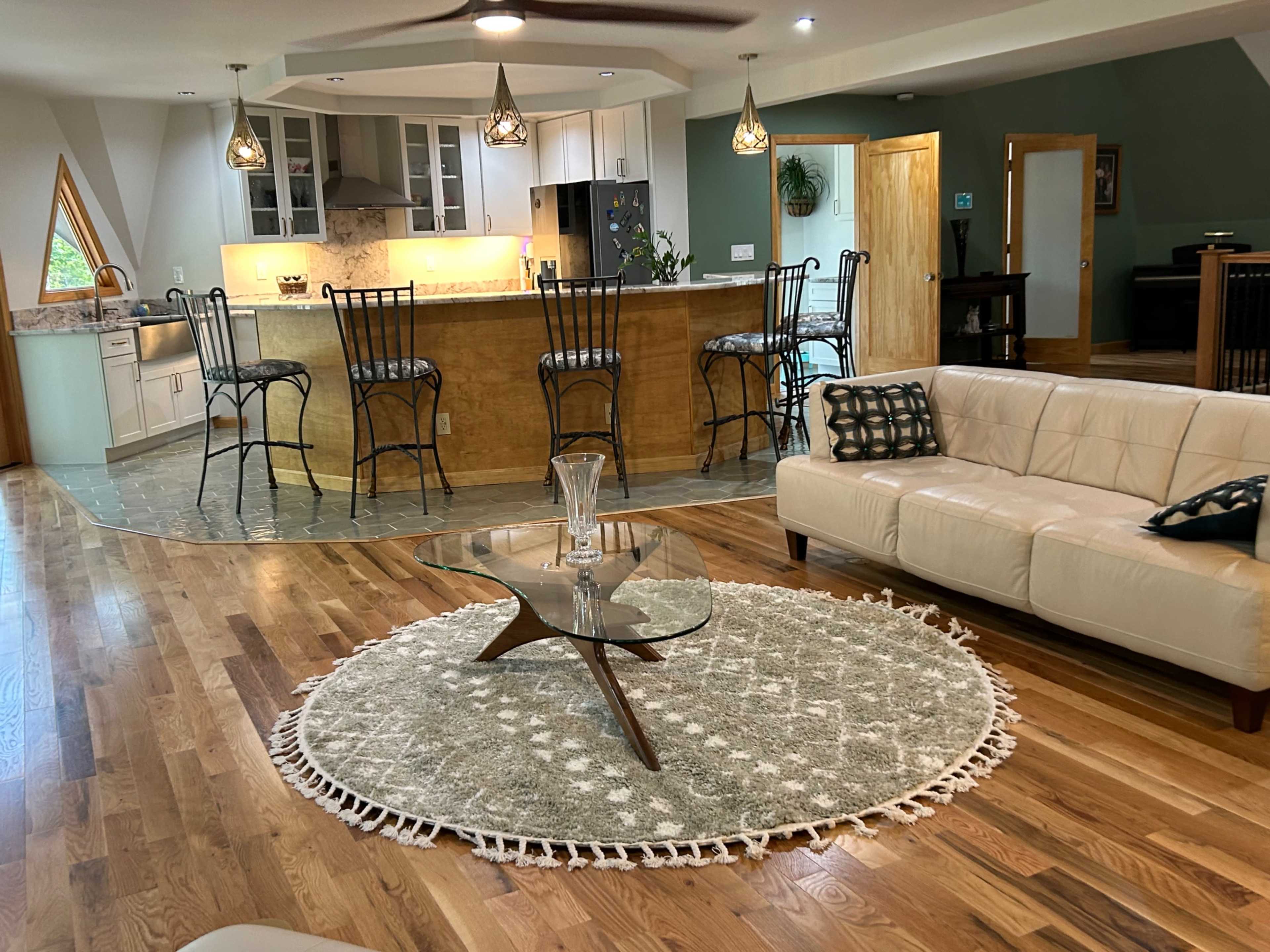 The image features a modern living area with a light-colored sofa, a round rug, a glass coffee table, and a kitchen with bar stools in the background.
