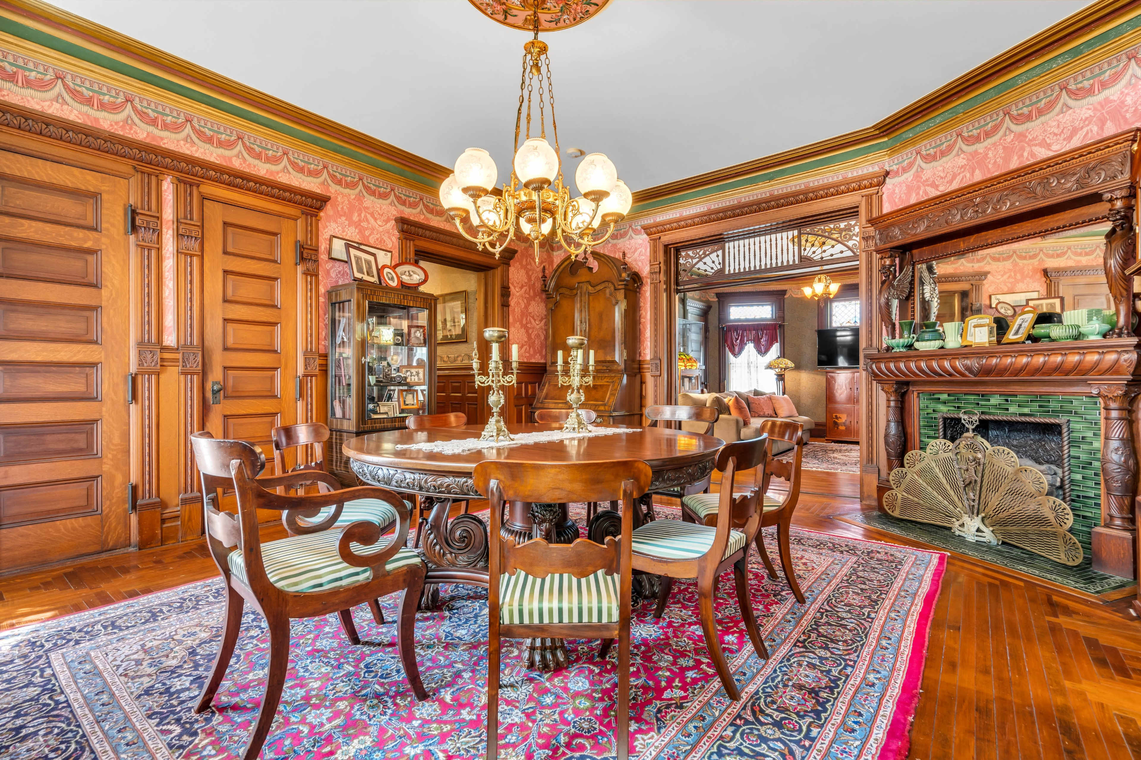 A ornate dining room with a round table, classic wooden chairs, and a chandelier, featuring patterned wallpaper and detailed woodwork.