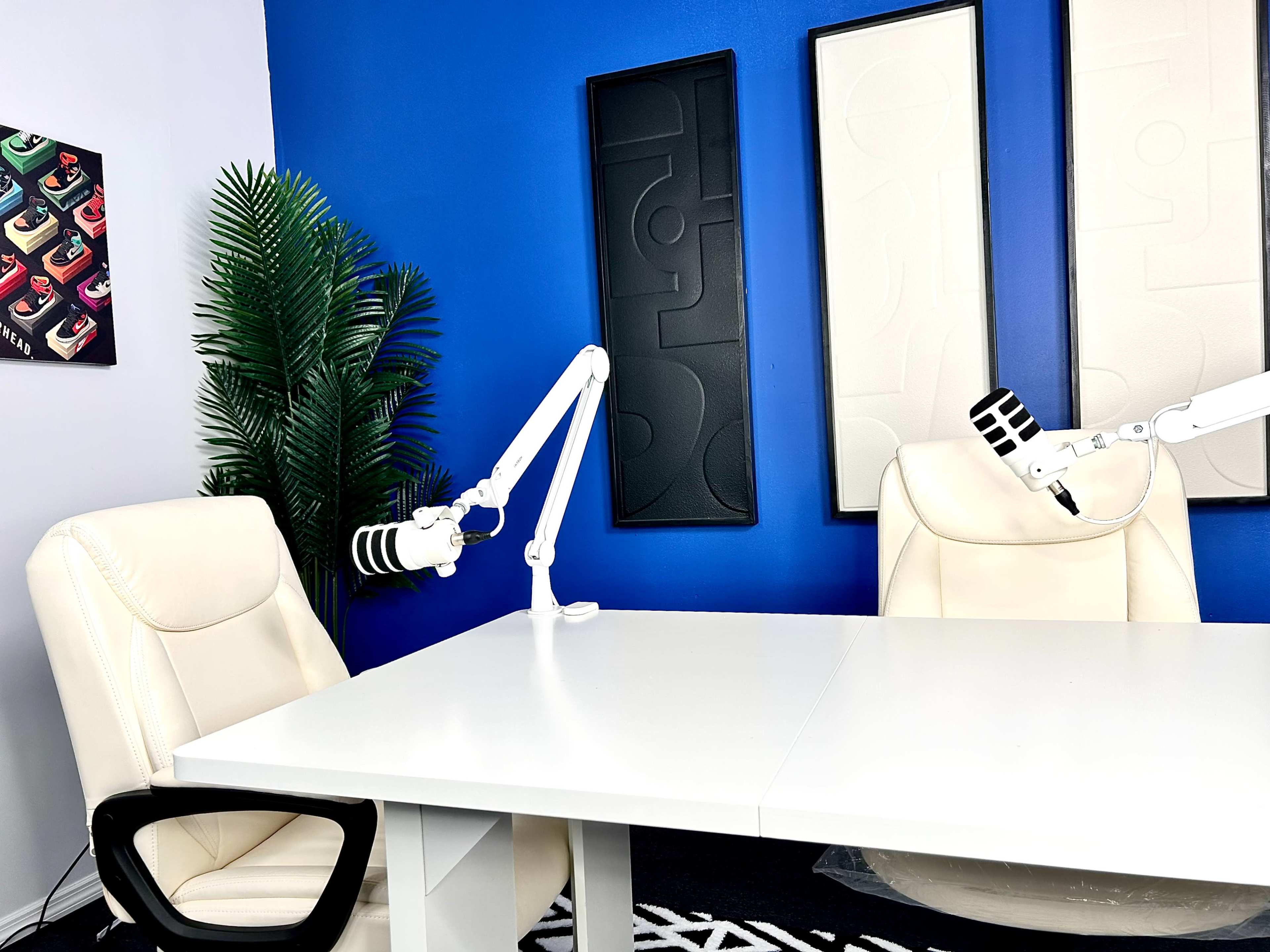 The image shows a bright studio space with two empty white chairs and adjustable microphones mounted on a white table, against a blue wall with abstract art.