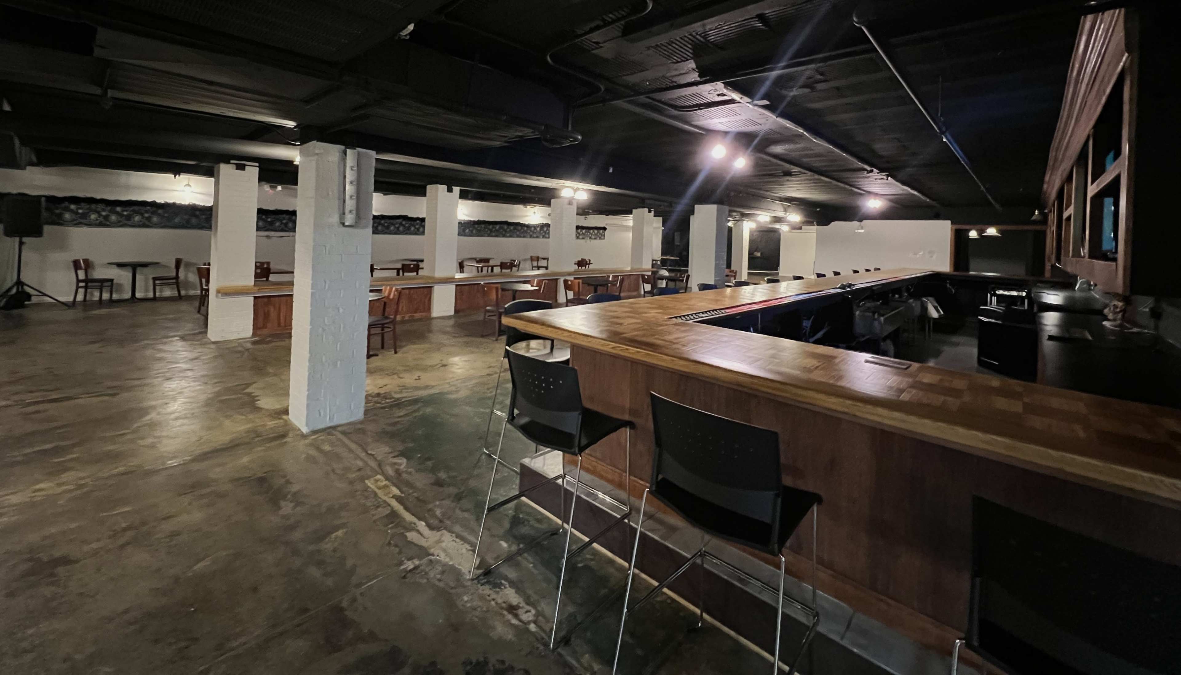 A spacious, dimly lit bar area with empty tables and a long wooden counter.