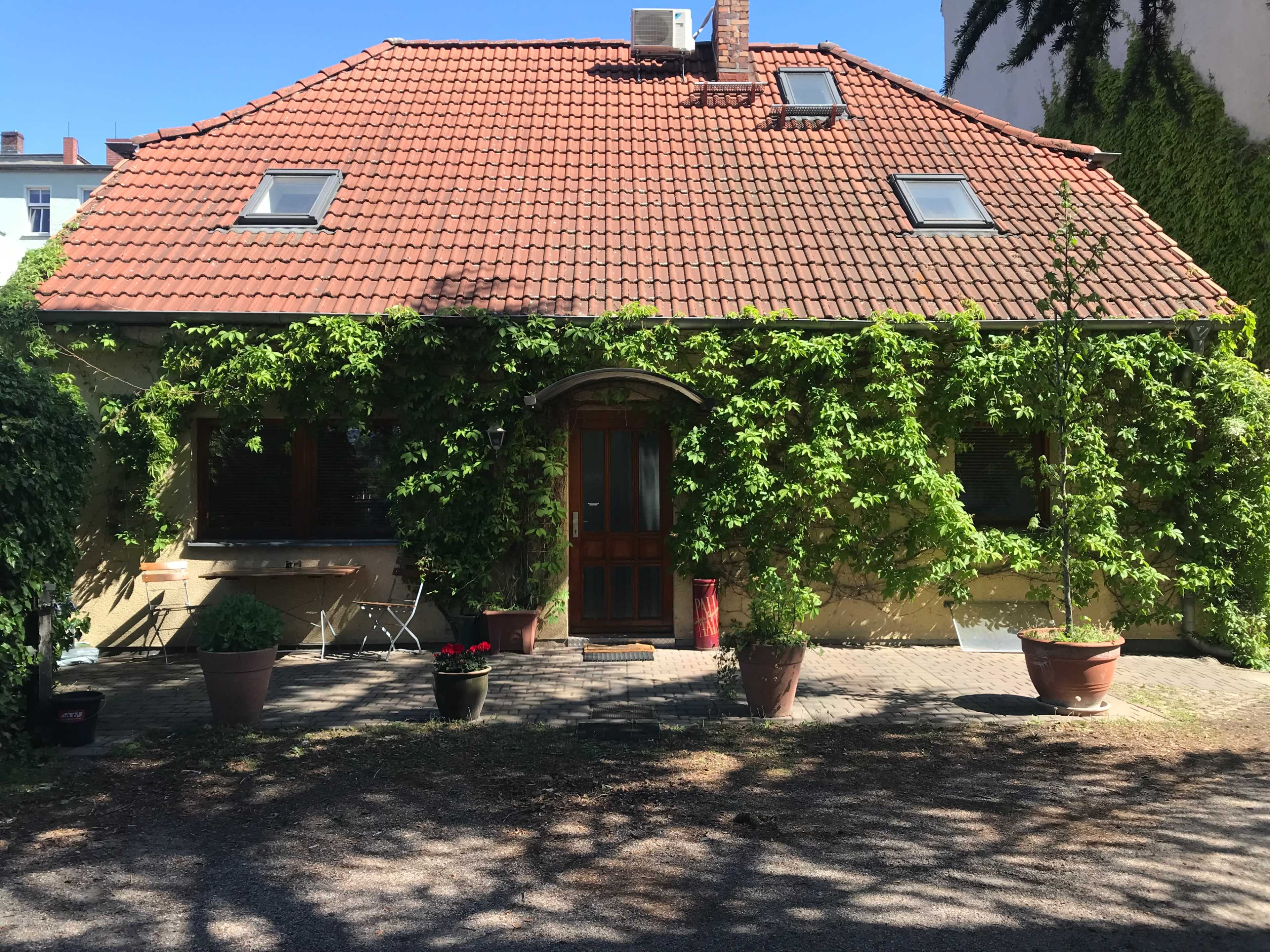 The image shows a house with a red tiled roof, surrounded by green vines, and a small garden area featuring potted plants.