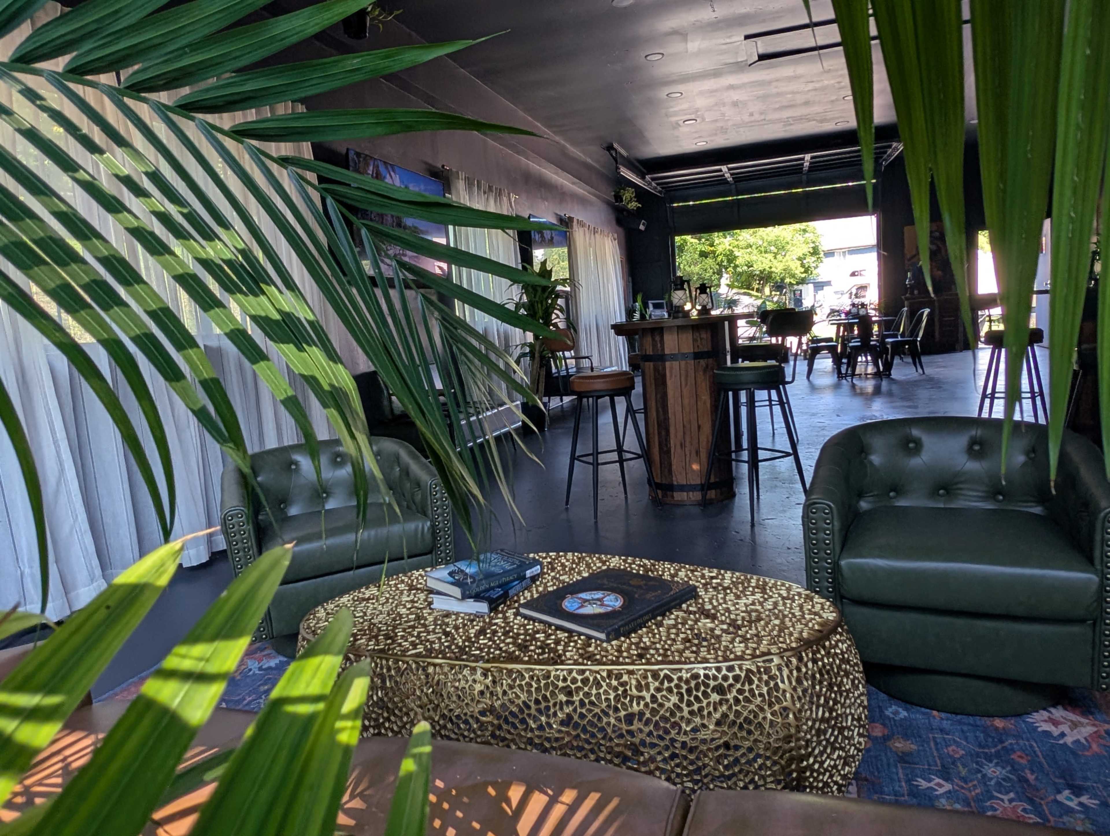 The image shows a stylish, open lounge area with green seating, a decorative table, and high tables in the background surrounded by plants.