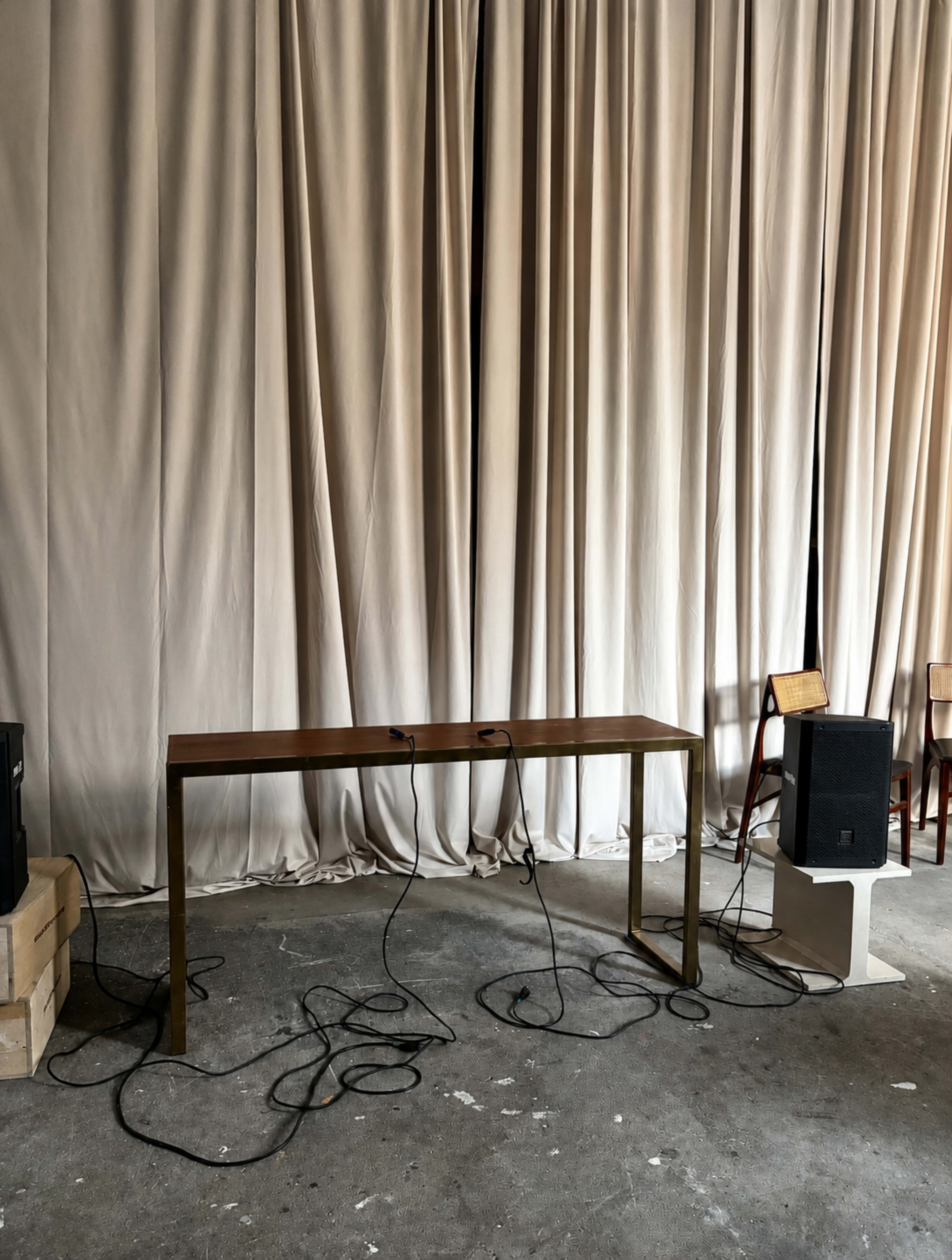 A long, minimalistic table with tangled cables is positioned in front of a backdrop of soft, draped curtains in a sparse room.