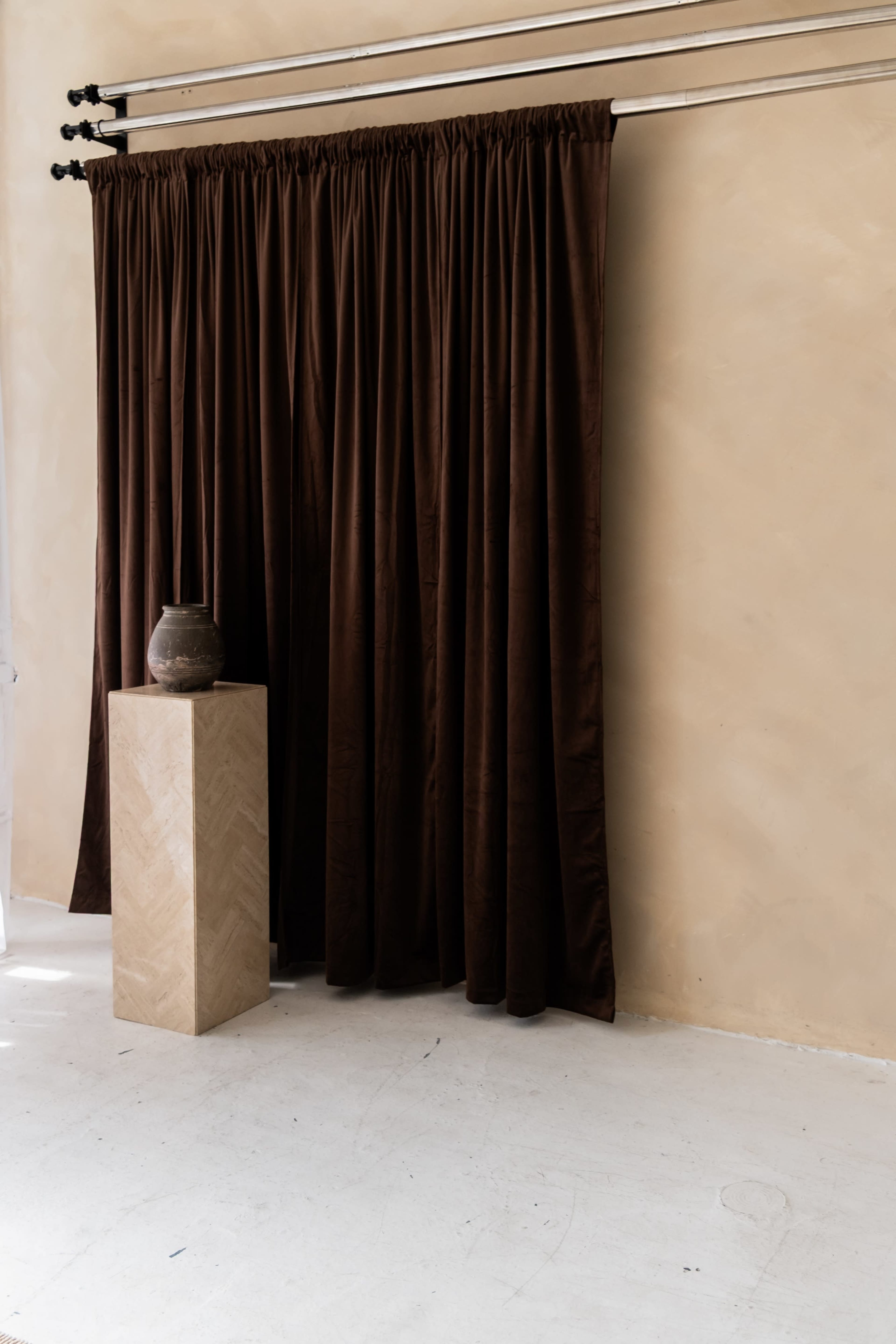 The image features a corner of a room with a large brown curtain hanging from a metal track, alongside a stone pedestal that holds a pottery vessel, set against a beige wall and white floor.