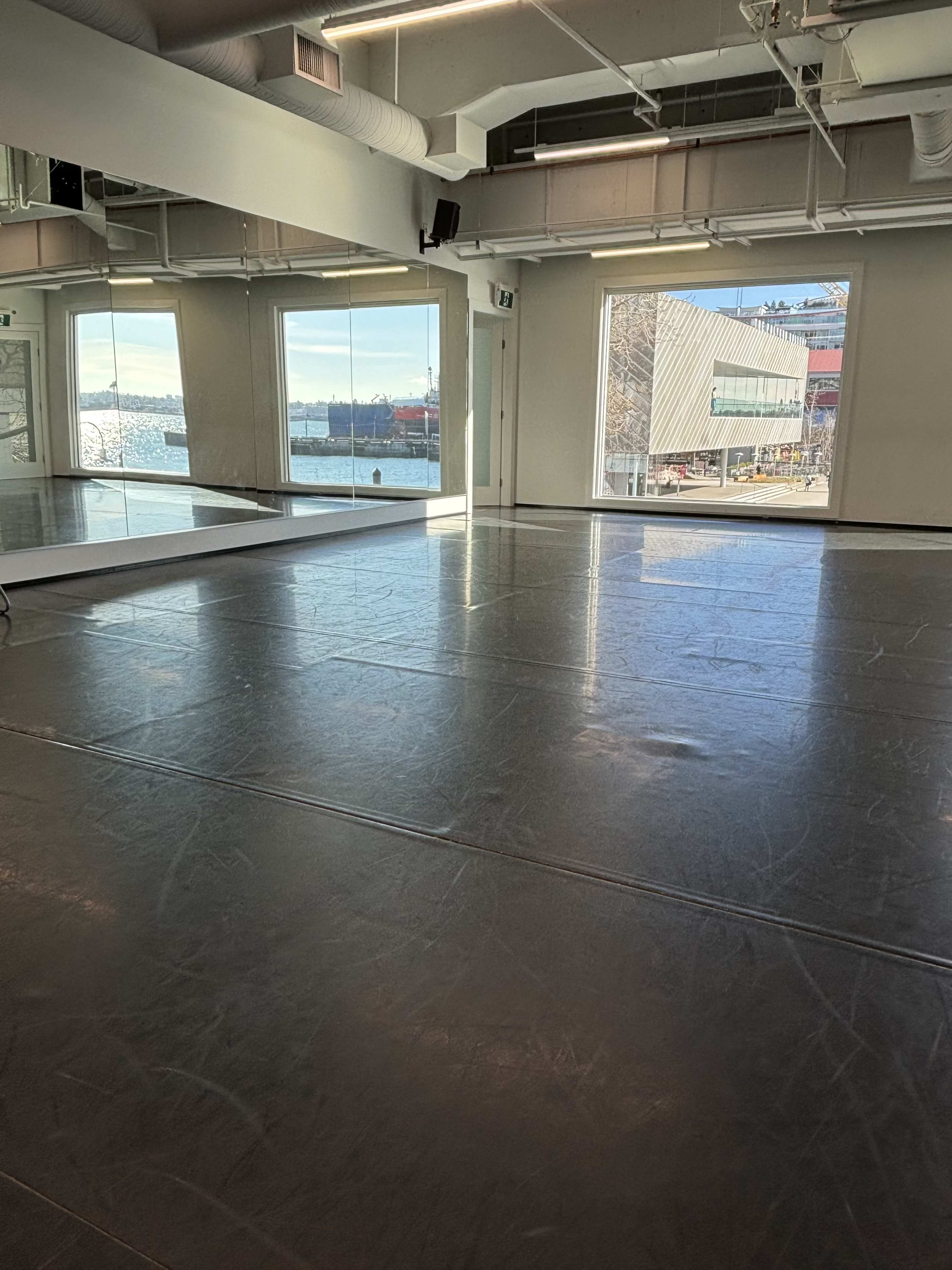 The image shows a spacious dance studio with large windows overlooking a waterfront scene.